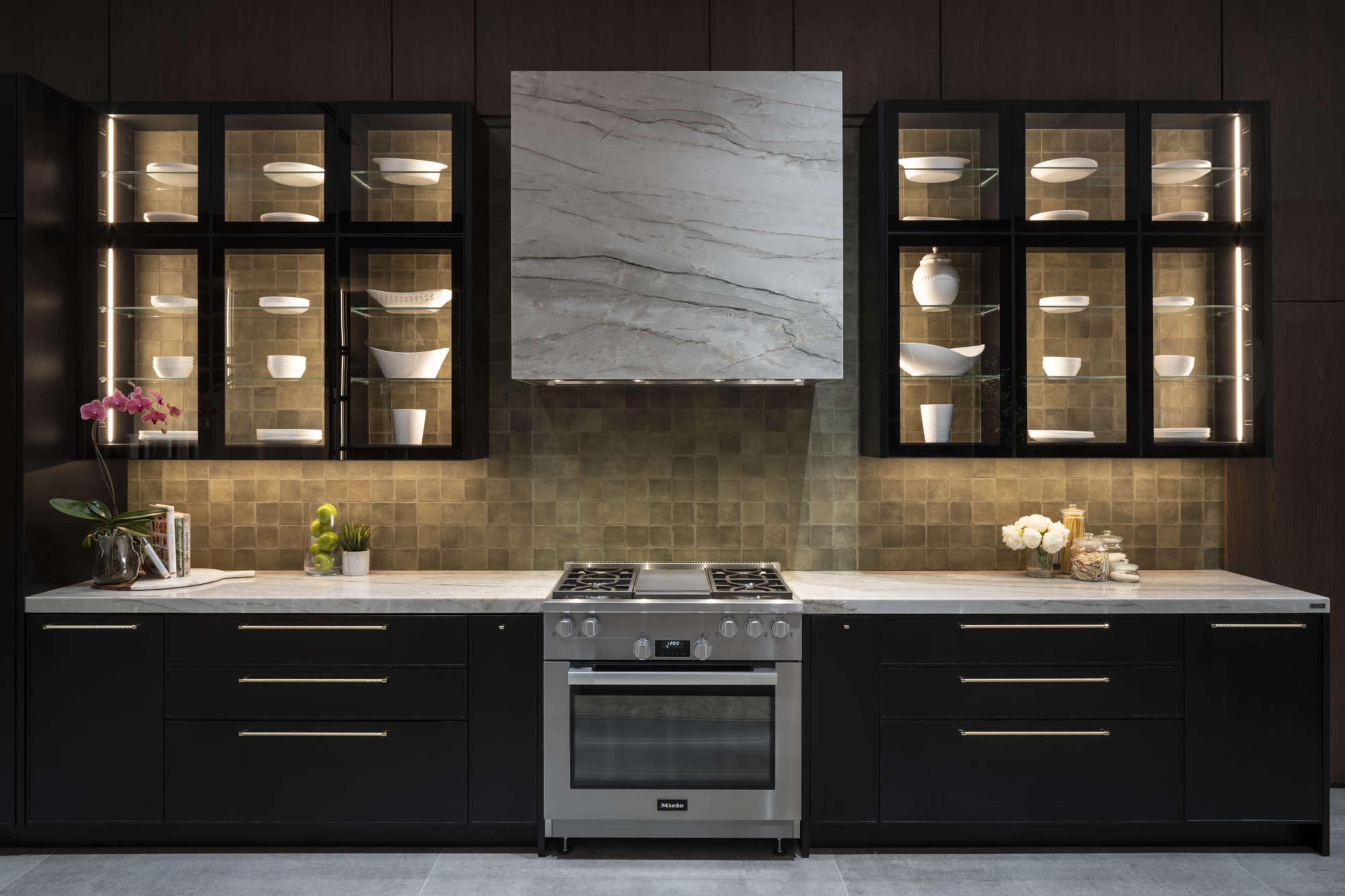 The image shows a modern kitchen with black cabinetry, a marble backsplash, and stainless steel appliances, featuring glass-front cabinets displaying white dishware.