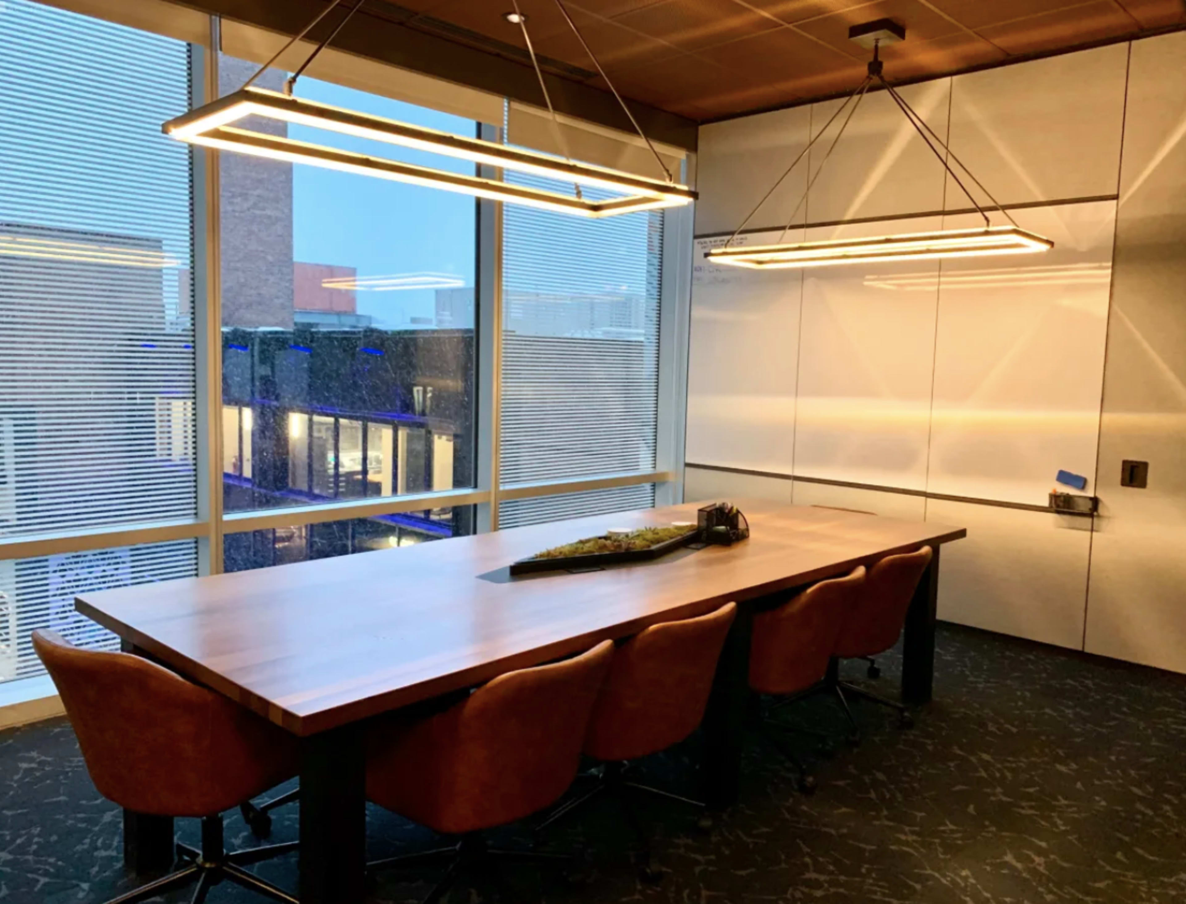 A modern conference room features a large table surrounded by brown upholstered chairs and illuminated by two linear pendant lights.
