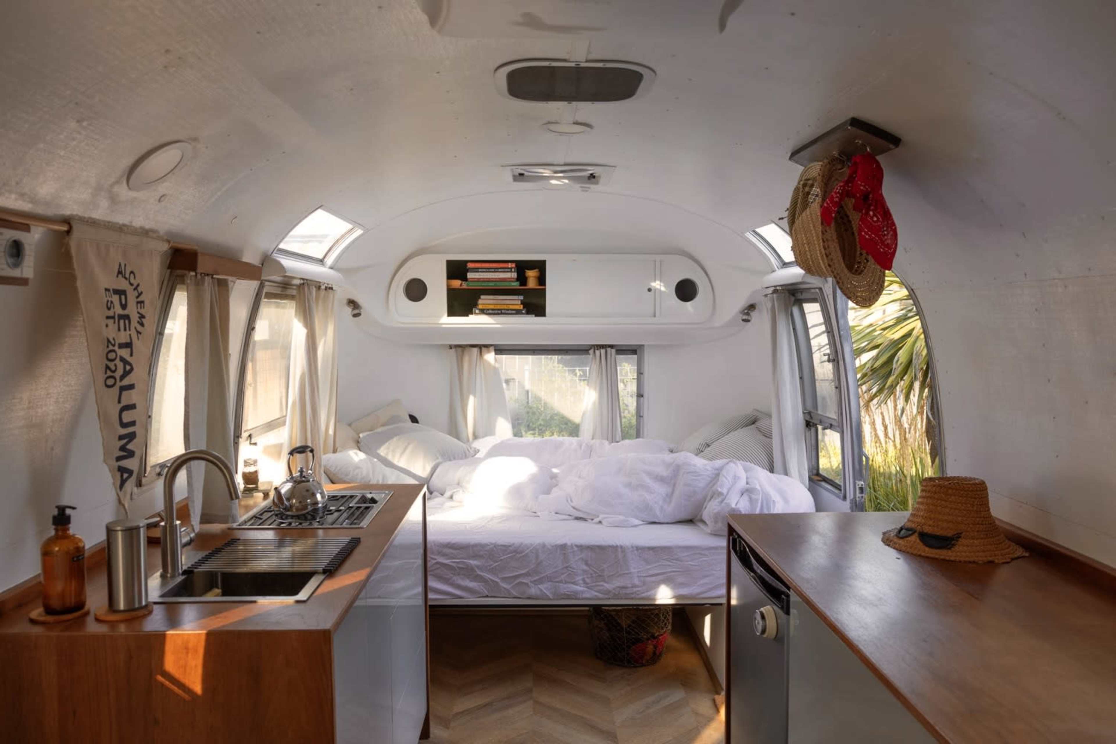 The interior of a streamlined Airstream trailer features a spacious sleeping area with a bed, small kitchen, and wooden cabinetry, illuminated by natural light from overhead windows.