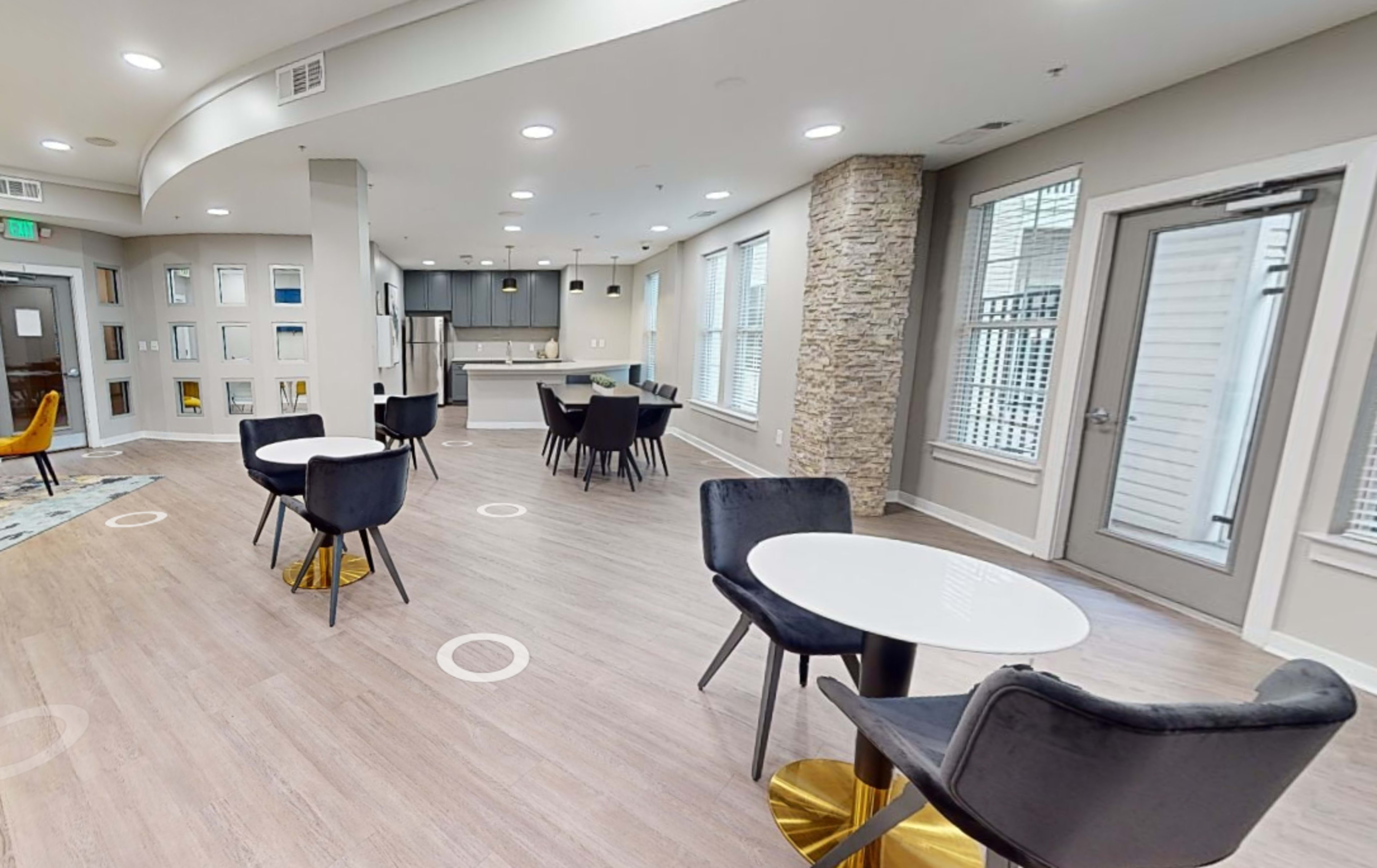 A modern lounge area with seating arrangements, including tables and chairs, and a kitchenette in the background.