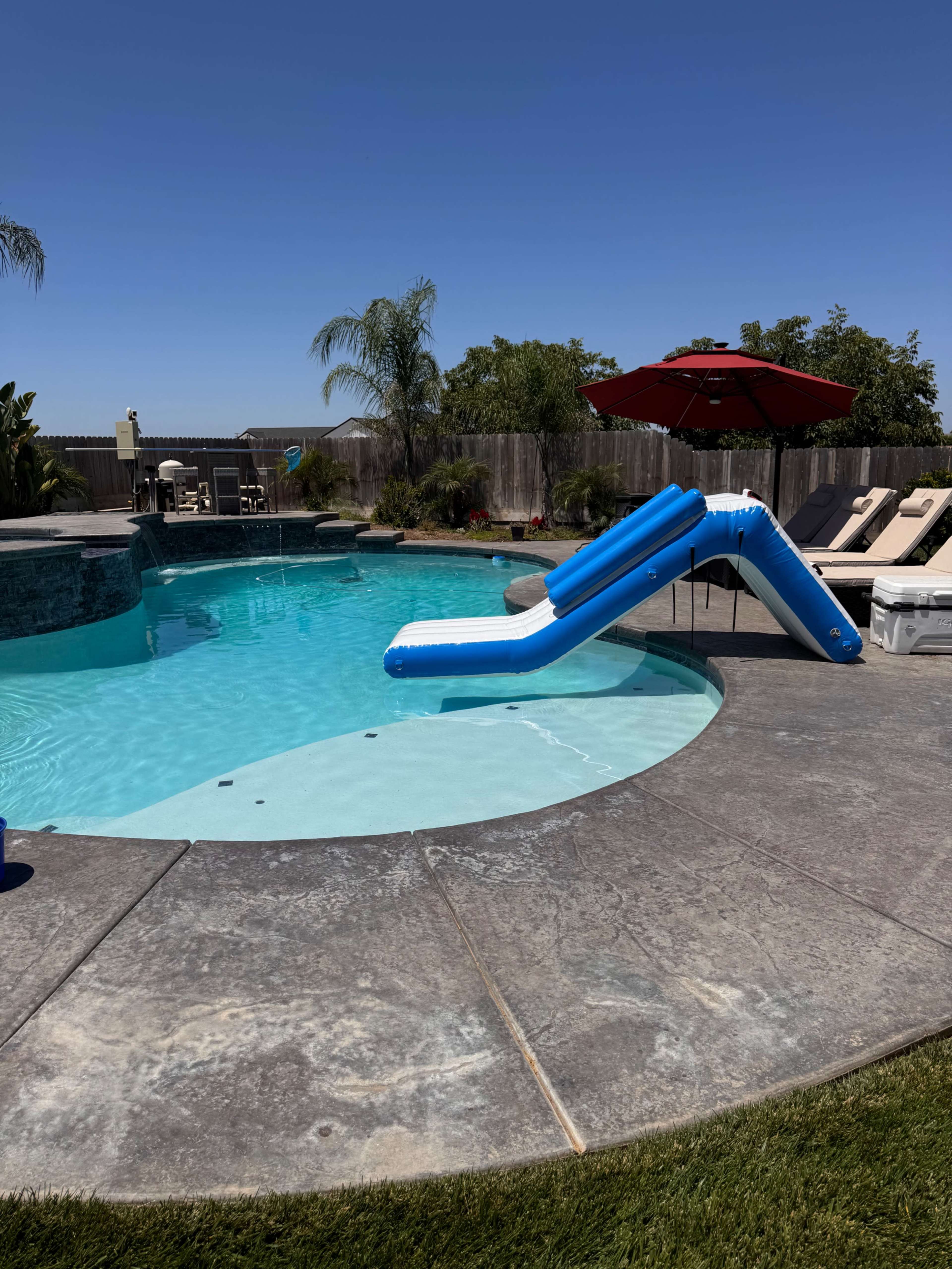 The image shows a swimming pool with a blue water slide and a red umbrella, surrounded by lounge chairs and greenery.