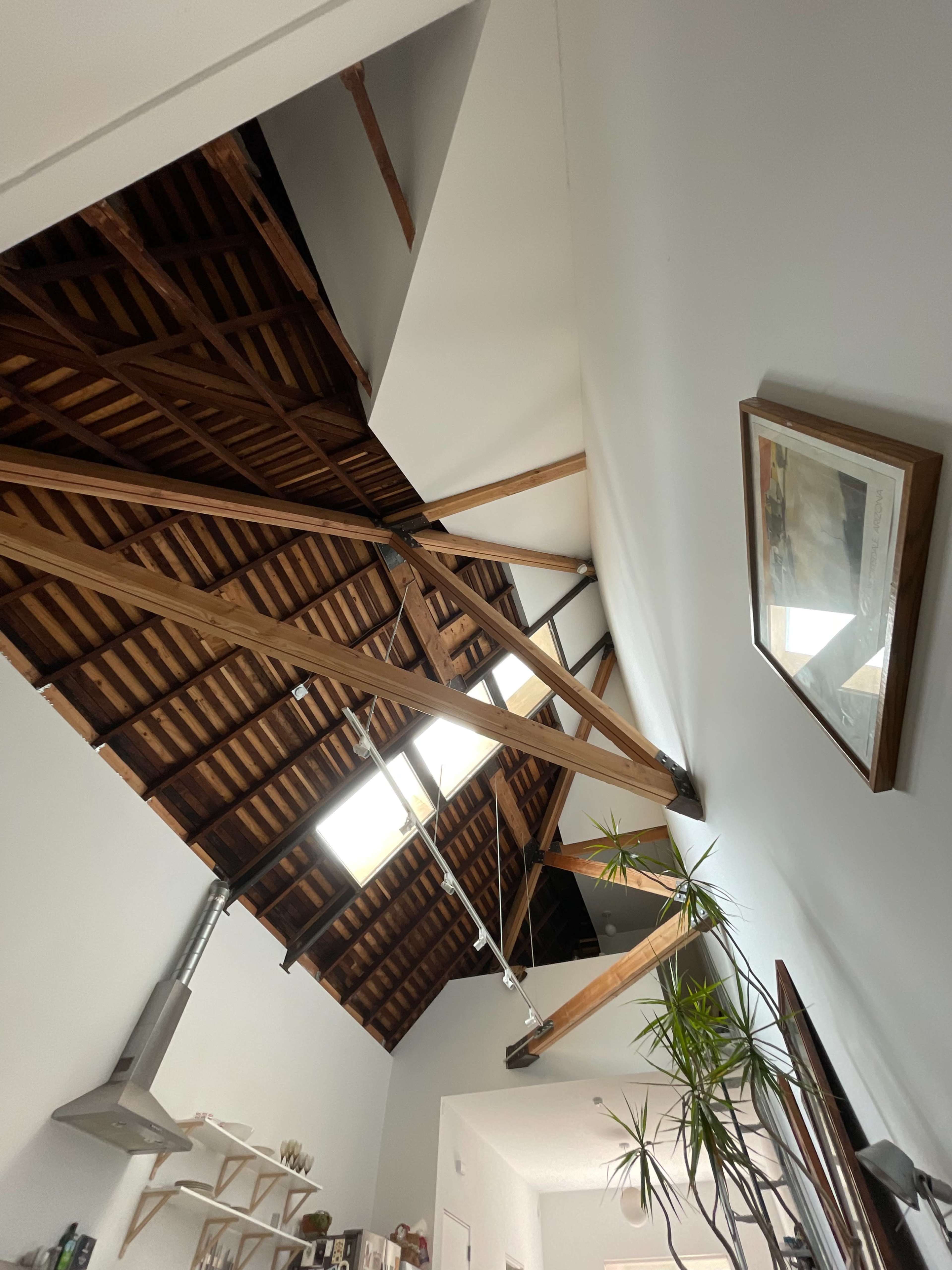 The image shows the interior of a room with a high ceiling featuring exposed wooden beams and a slanted roof, illuminated by skylights.
