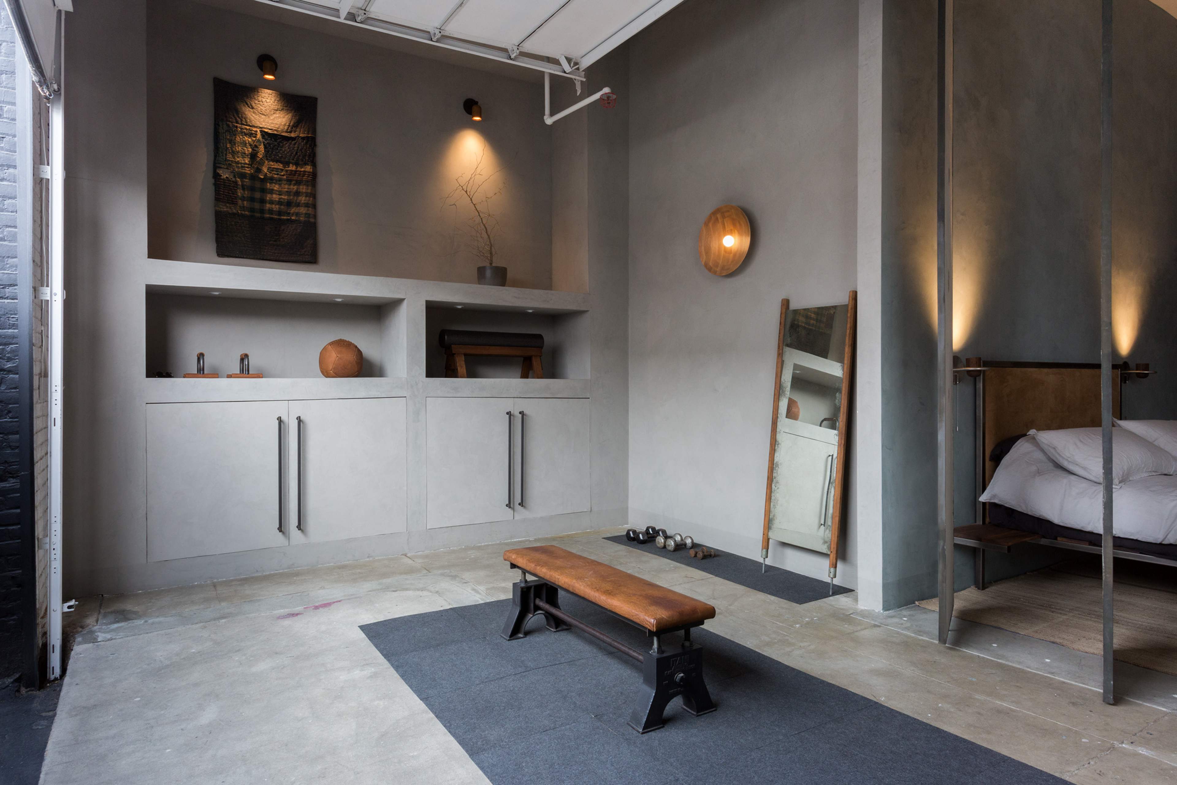 The image shows a minimalist interior of a garage-style gym with a wooden bench, small weights, a mirror, and shelves against a gray wall.
