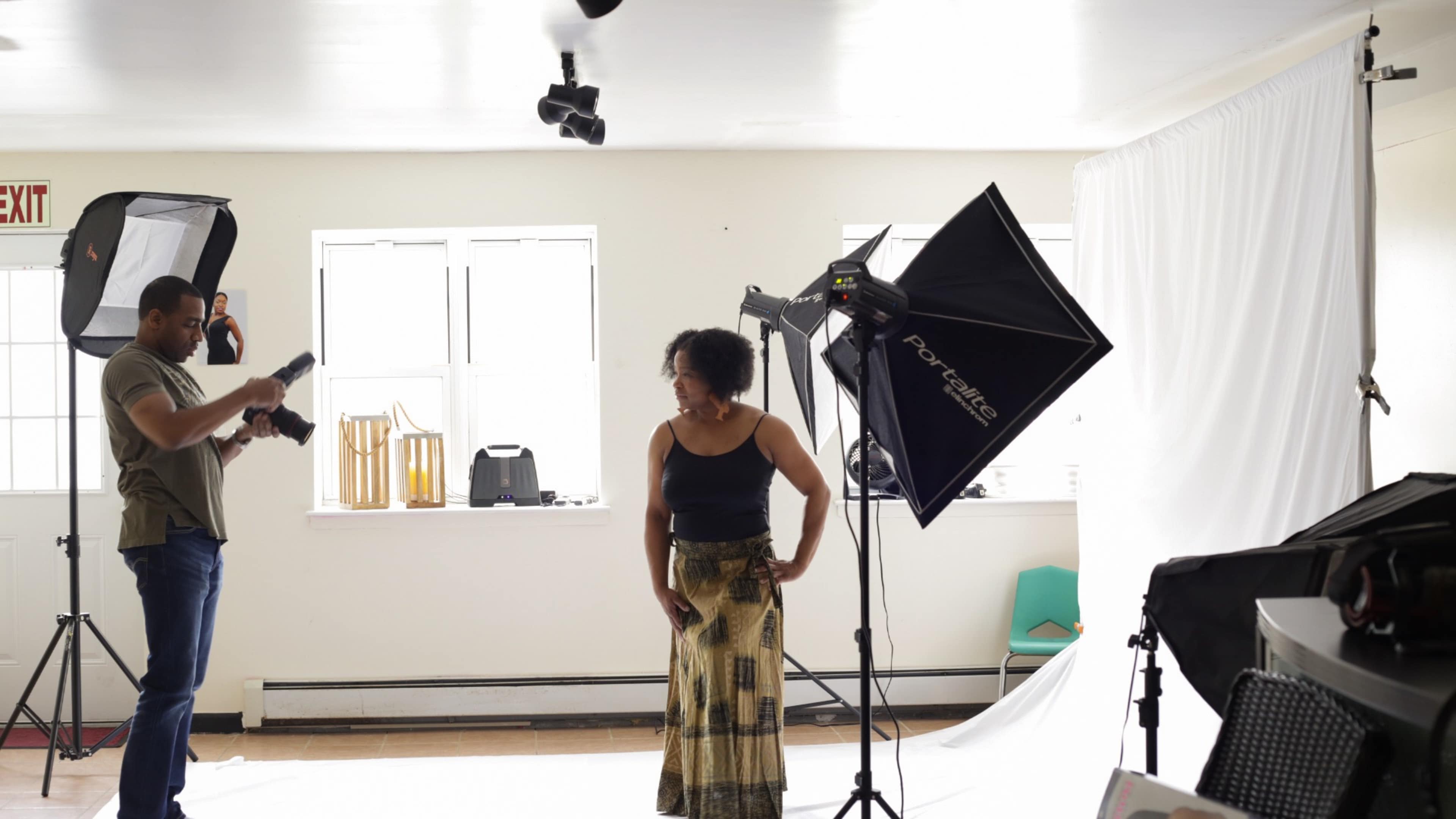A woman stands posing in a brightly lit photography studio while a man adjusts his camera equipment.