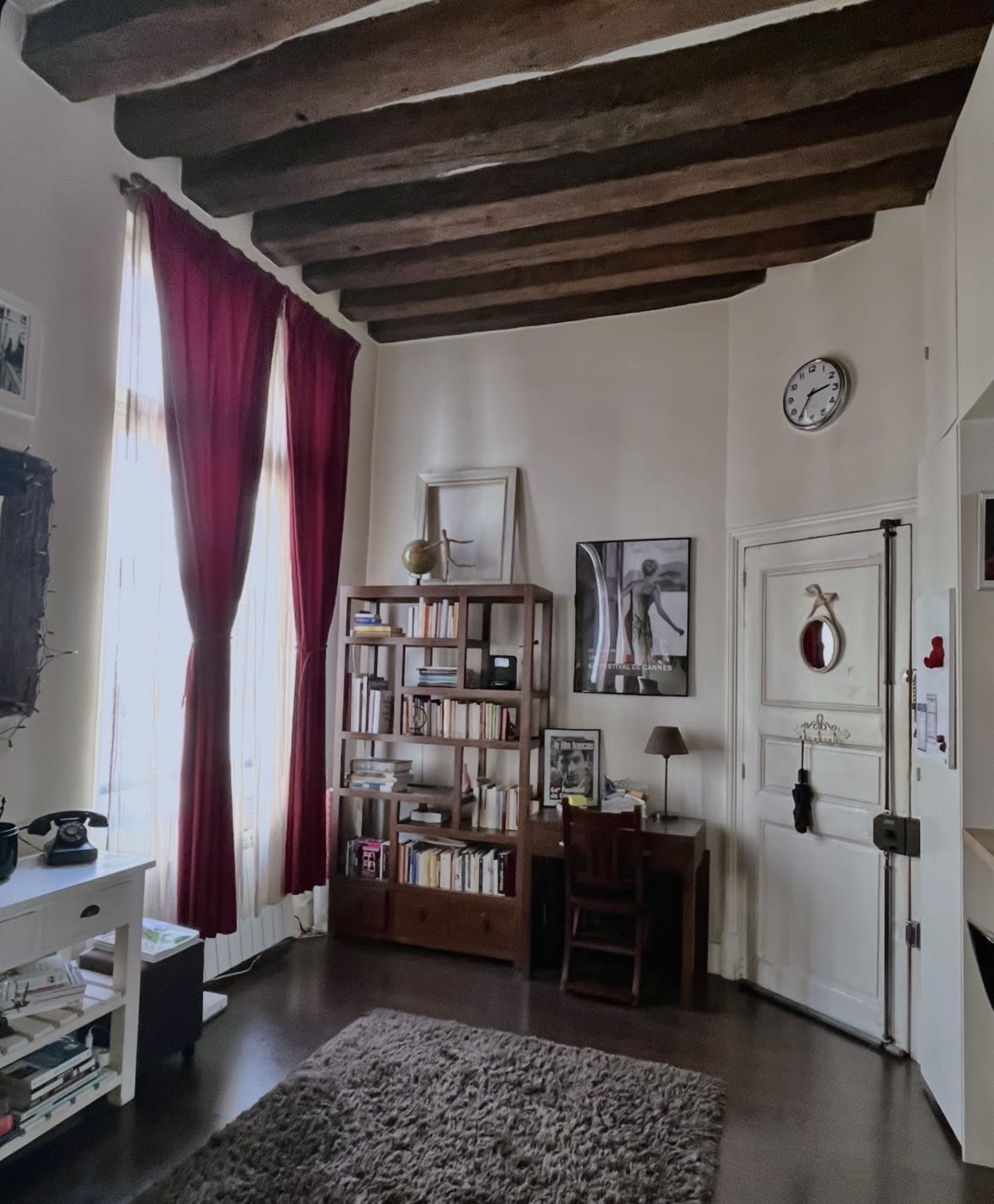 The image shows a cozy room featuring a bookshelf, a desk with a lamp, and large windows draped with red curtains.