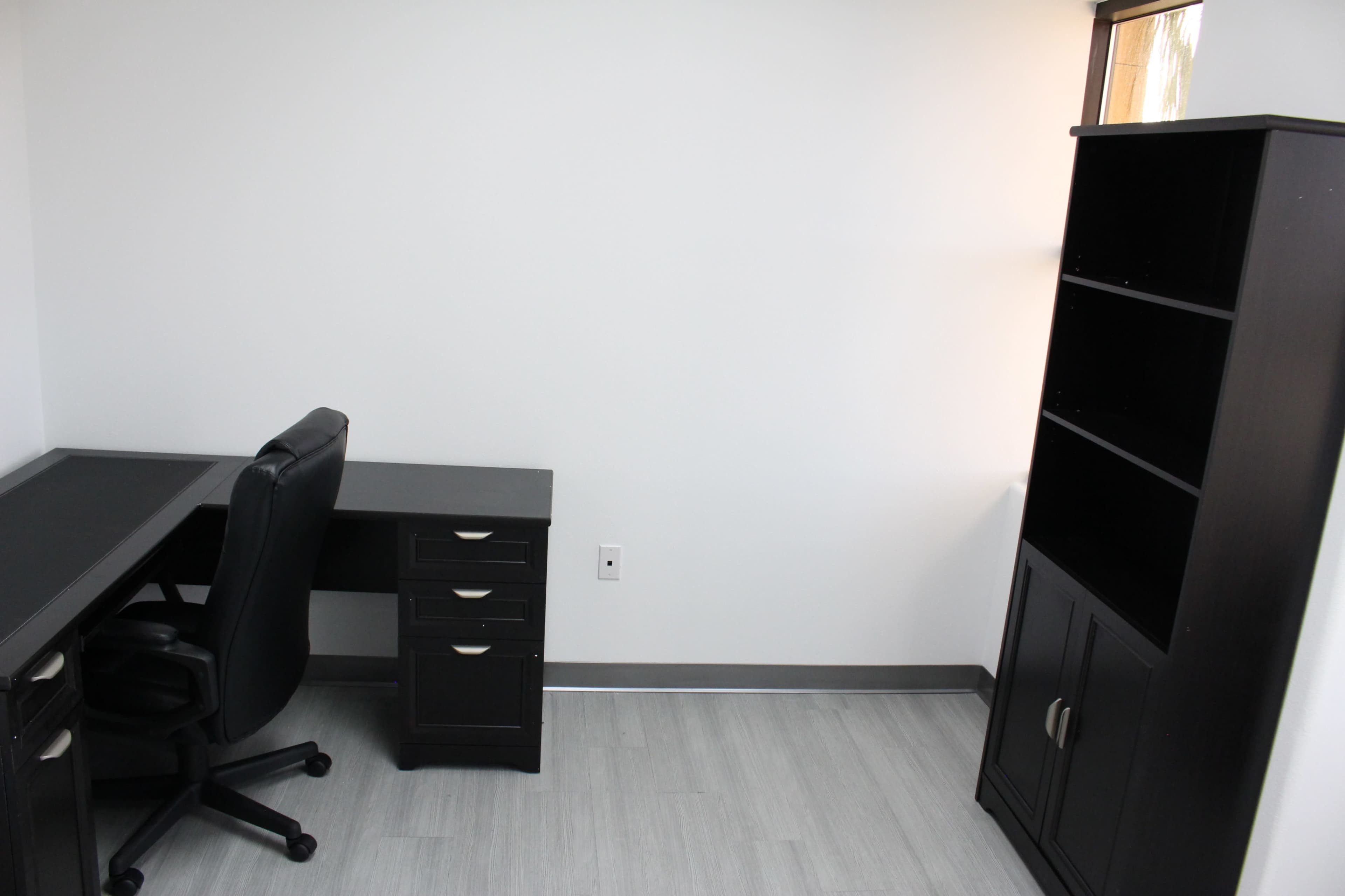 An empty office space featuring a black desk with drawers and a black office chair, alongside a tall shelf unit against a white wall.