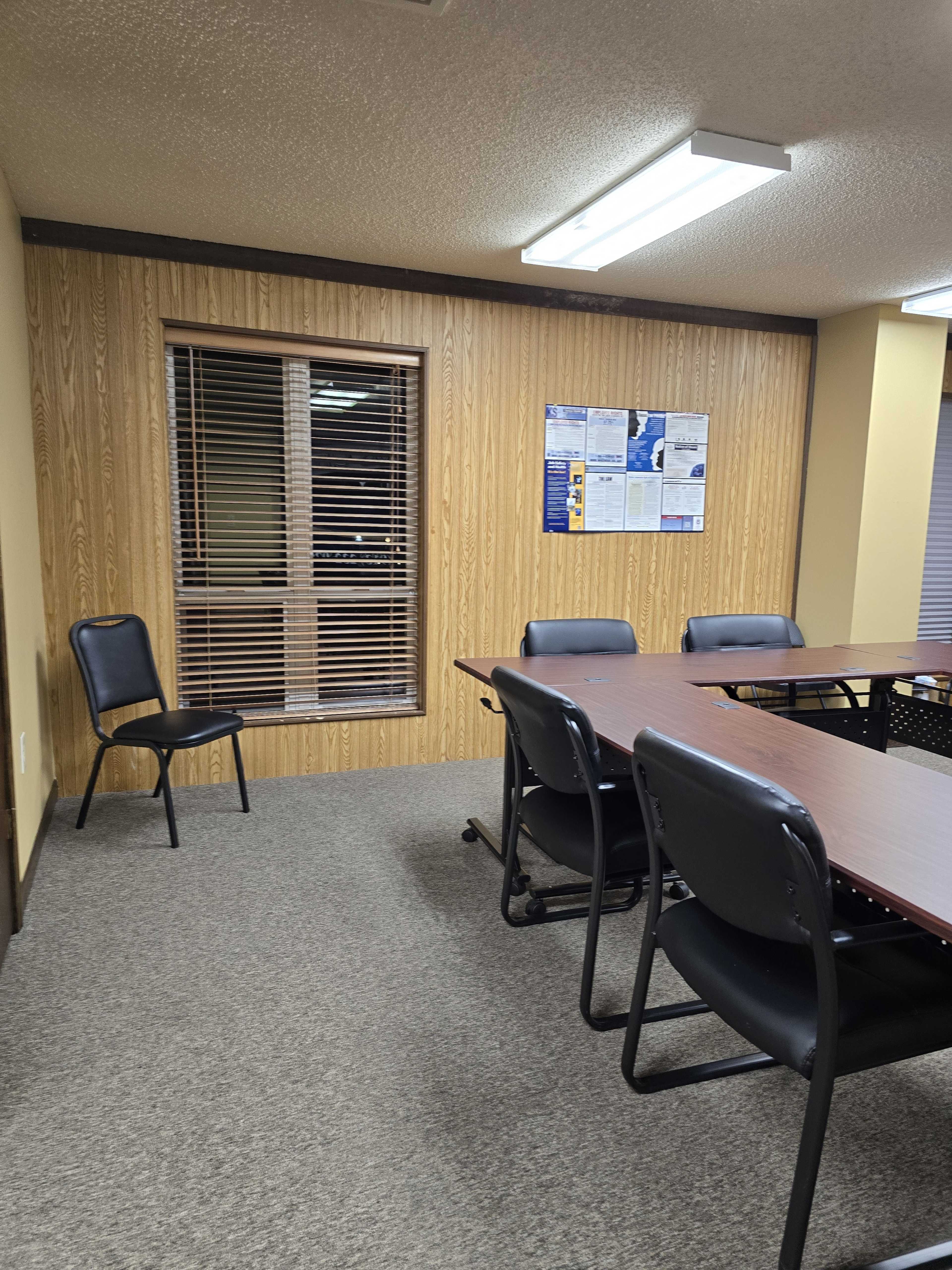 The image shows a conference room with a large table, several black chairs around it, and a window with blinds on one wall.