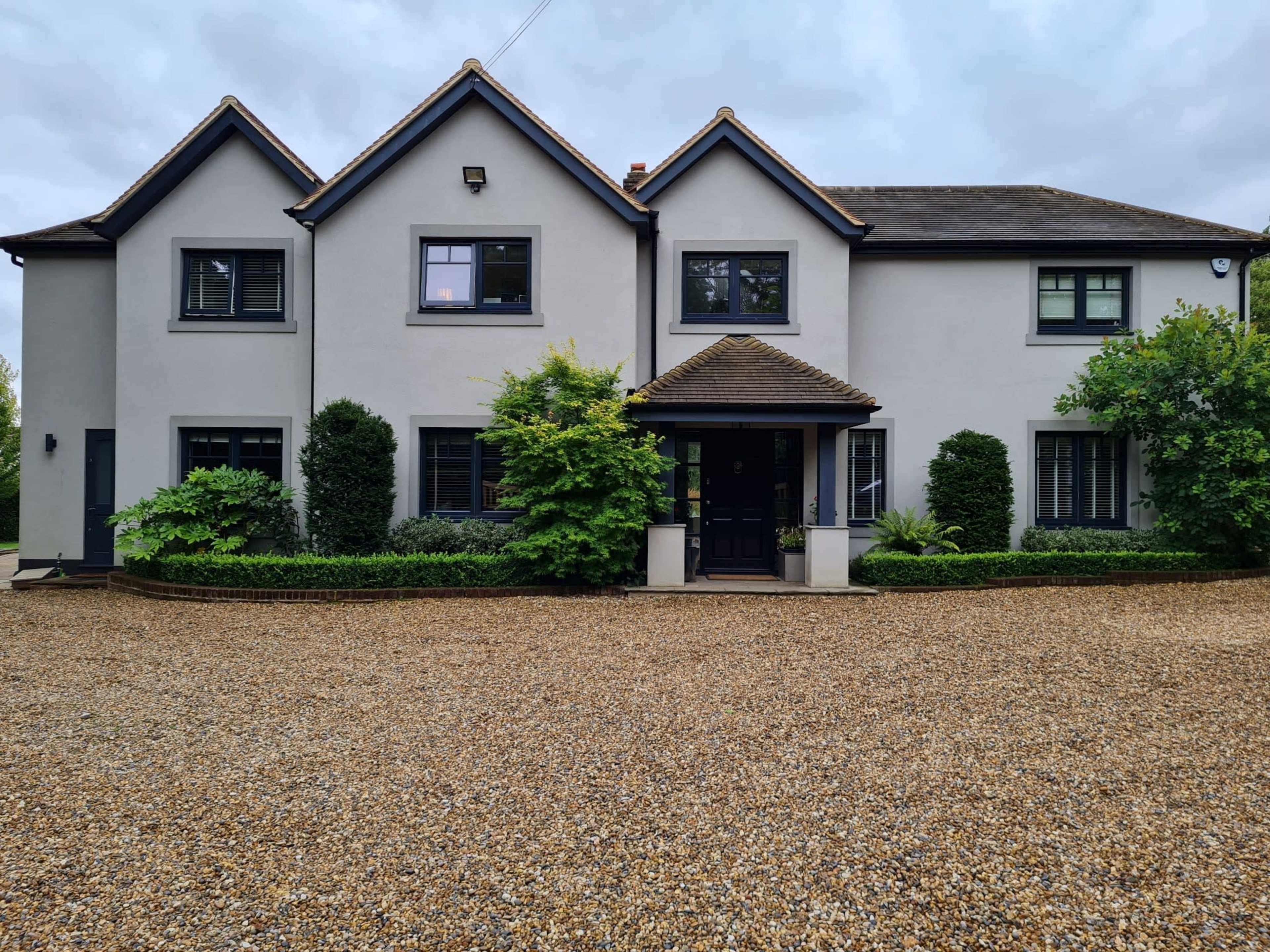 A modern two-story house with a gray exterior, a decorative entrance, and landscaped greenery surrounds a gravel driveway.