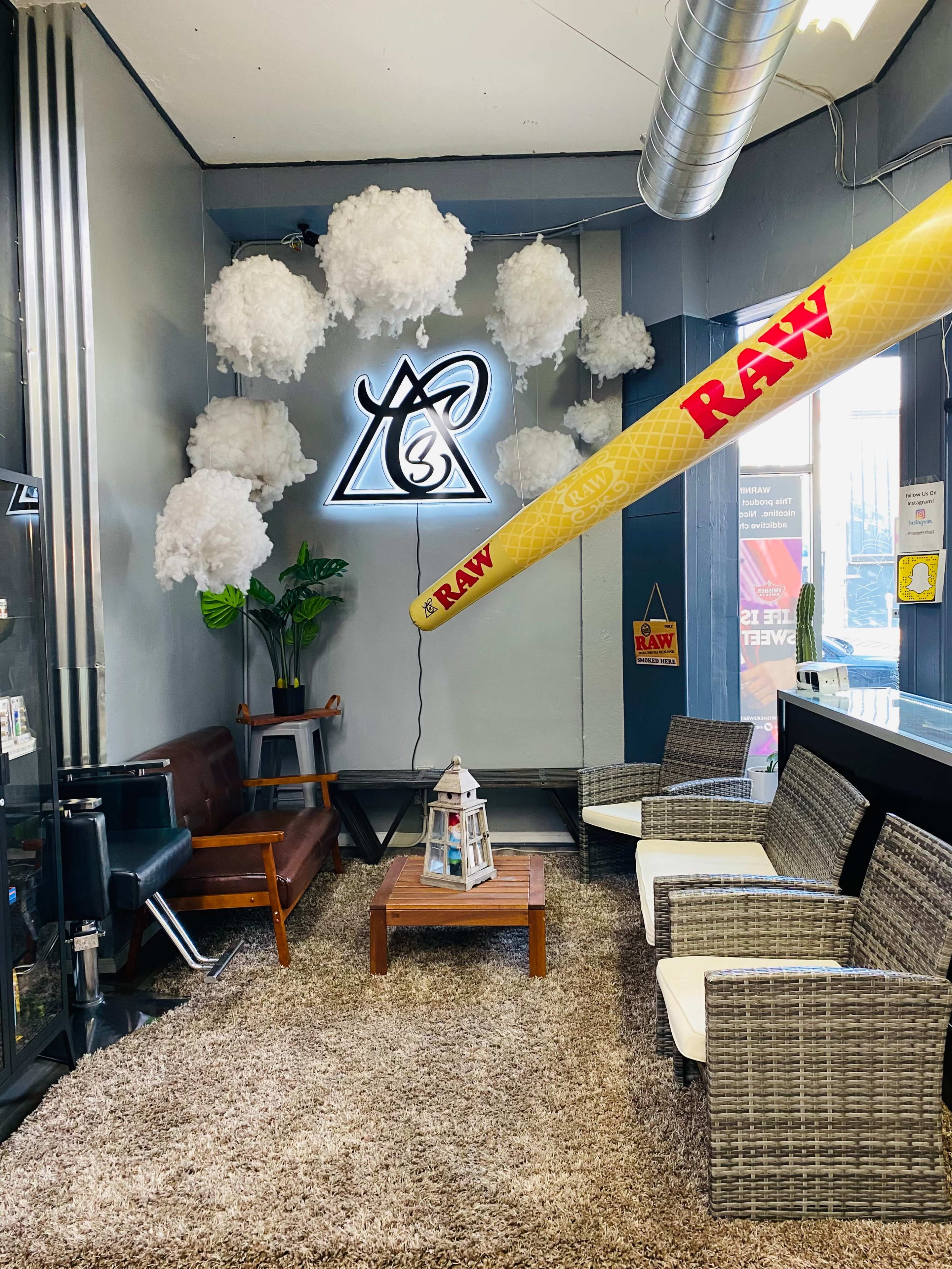 A lounge area features cloud-like decorations, a neon sign, wicker seating, a coffee table, and a large yellow RAW rolling paper prop.