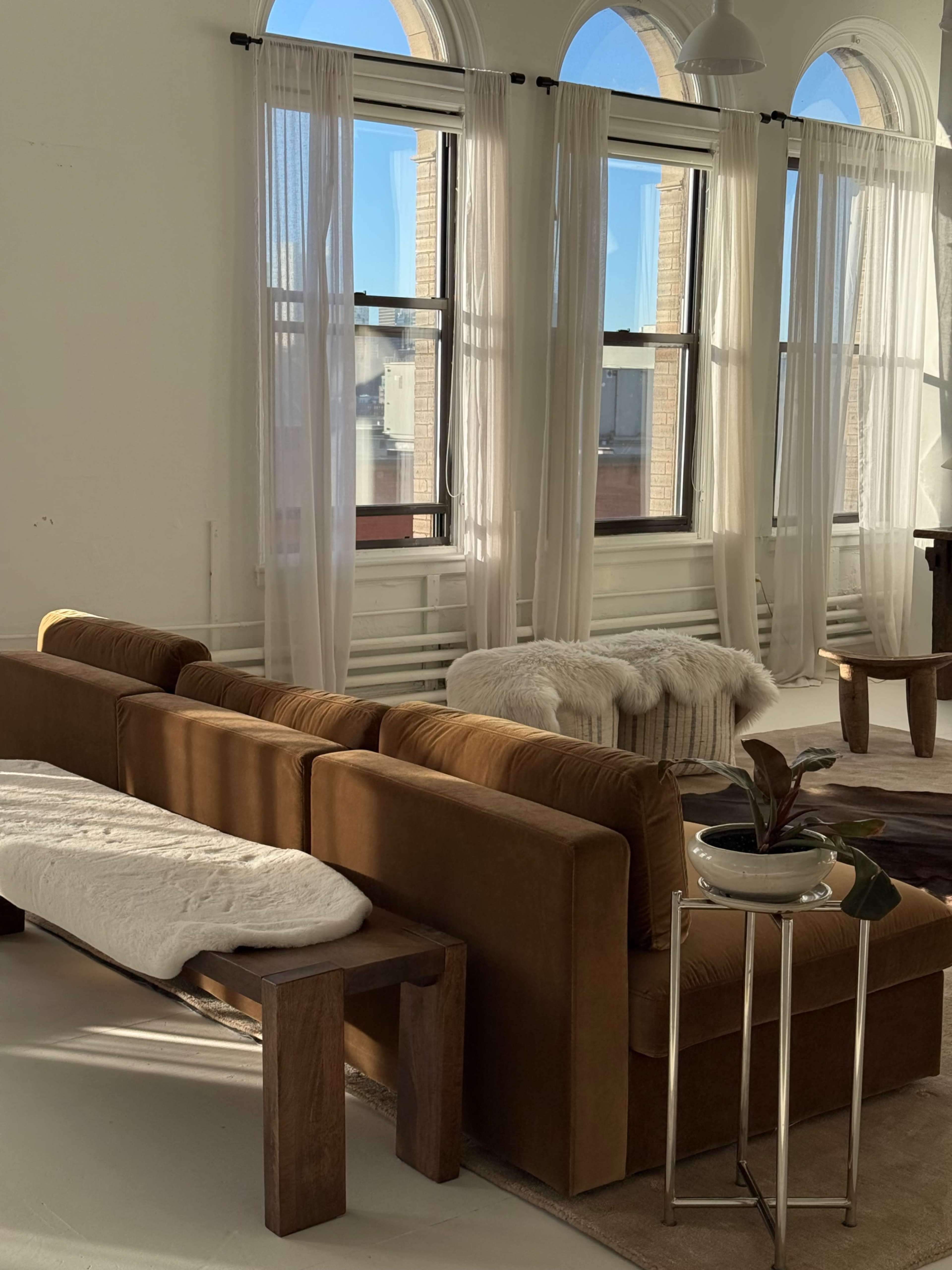 A modern living room features a brown sofa and a side table with a plant, illuminated by natural light from tall windows adorned with sheer curtains.