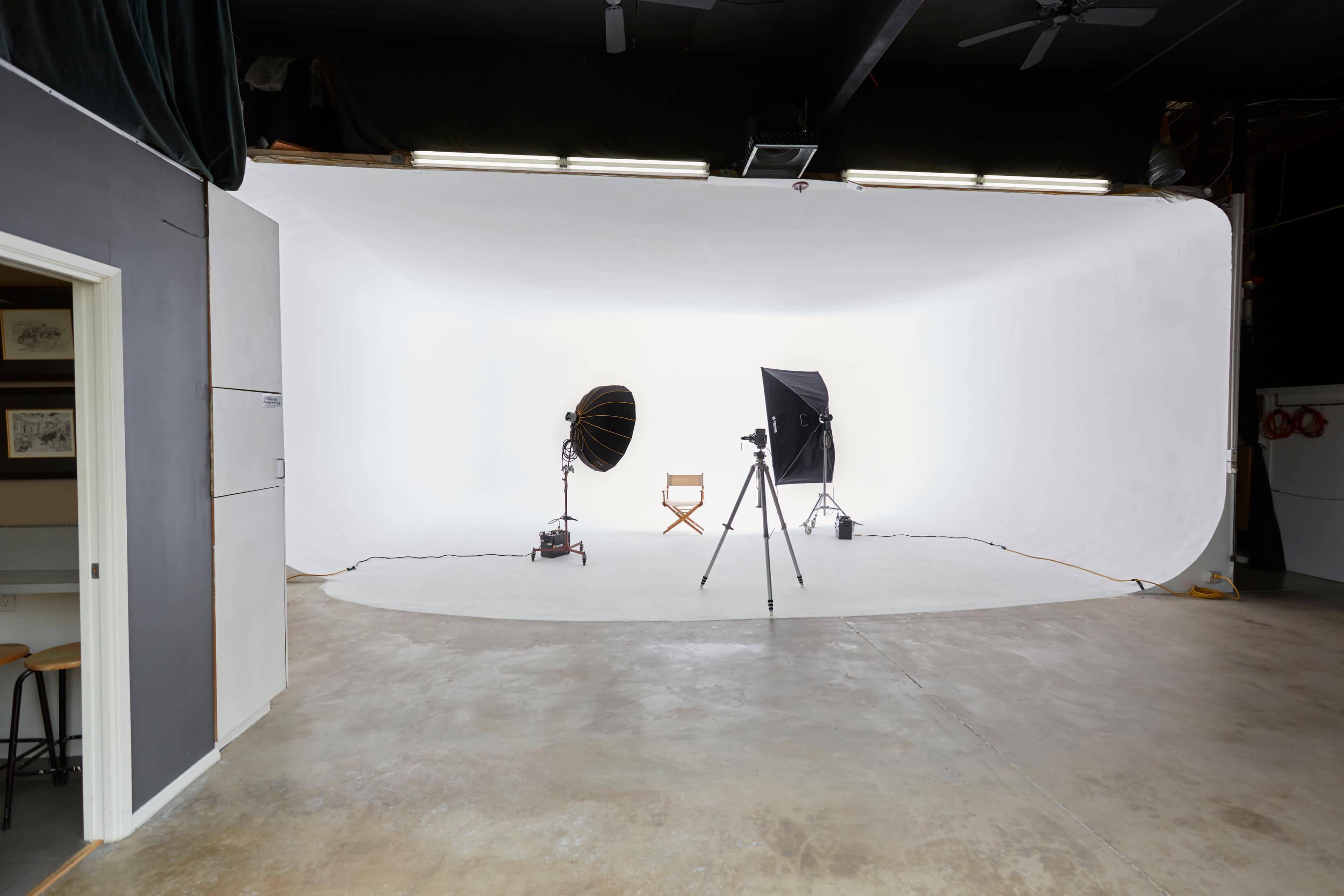 A photography studio features a seamless white backdrop, two light sources, a camera on a tripod, and a director's chair set up in the center.
