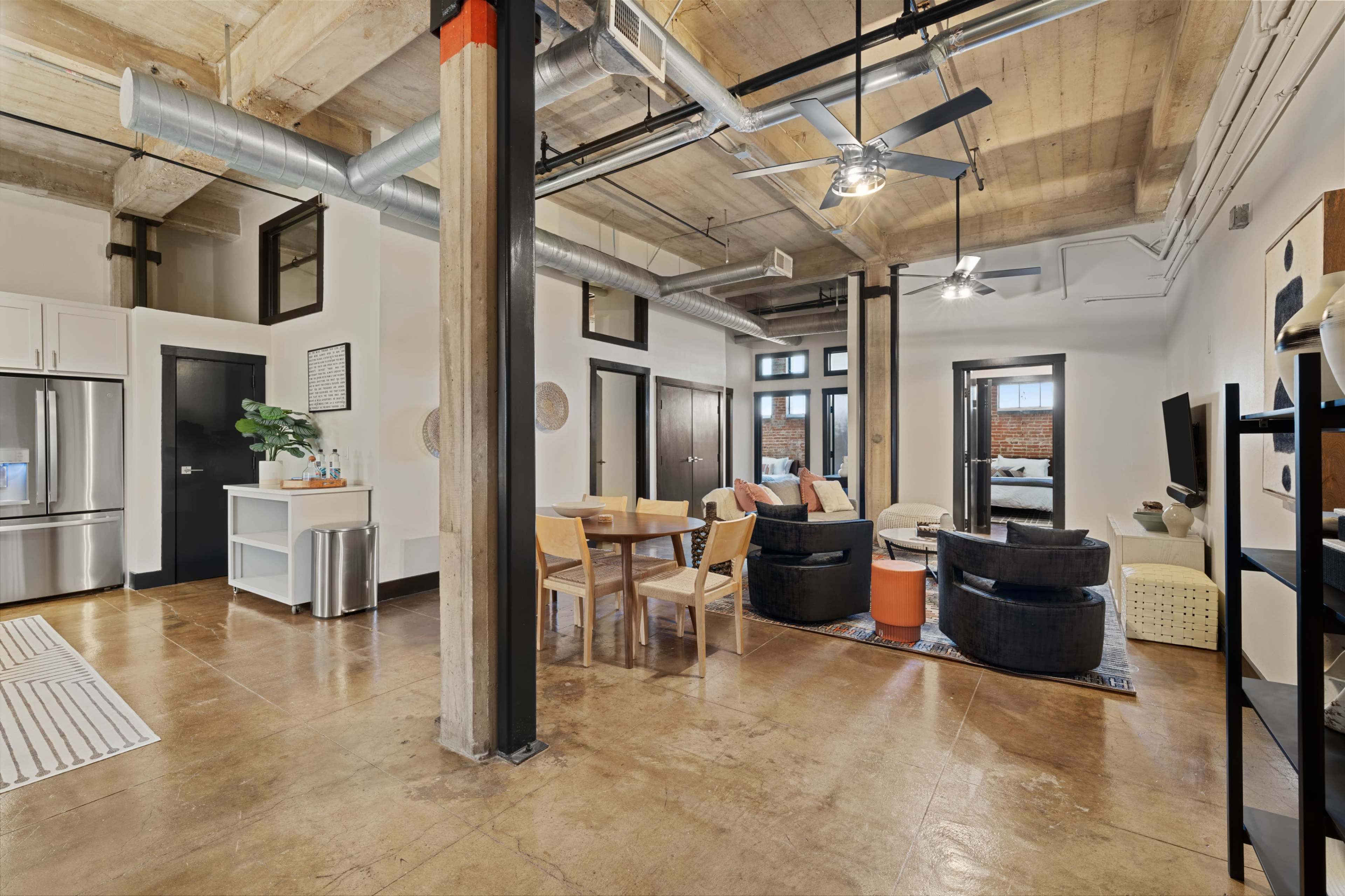 A modern loft-style living space featuring an open layout with a dining area, seating arrangement, and industrial-style elements such as exposed beams and ductwork.