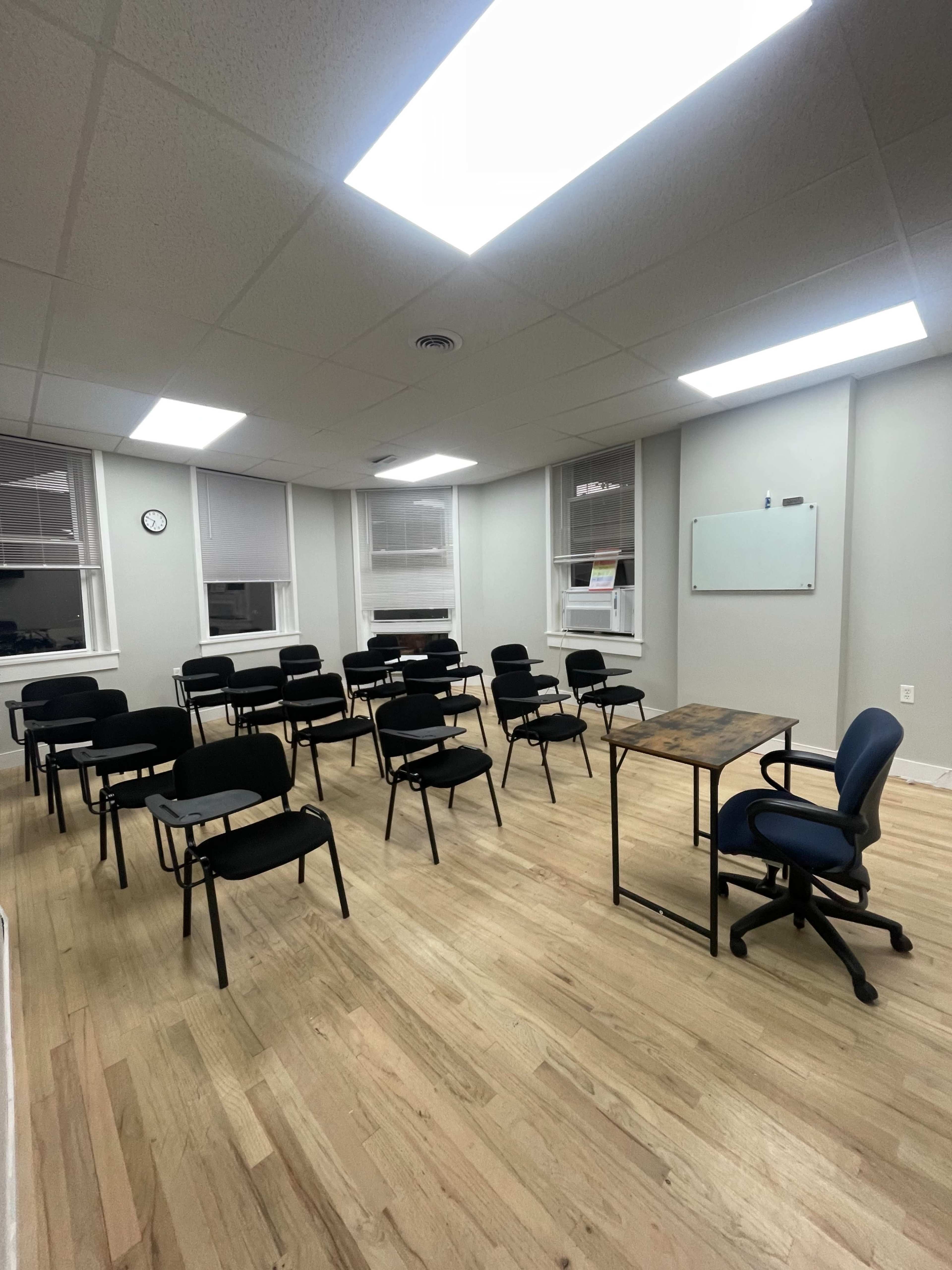The image shows a minimalist classroom setting with several rows of empty black chairs, a desk with a blue chair, and large windows allowing natural light to enter.
