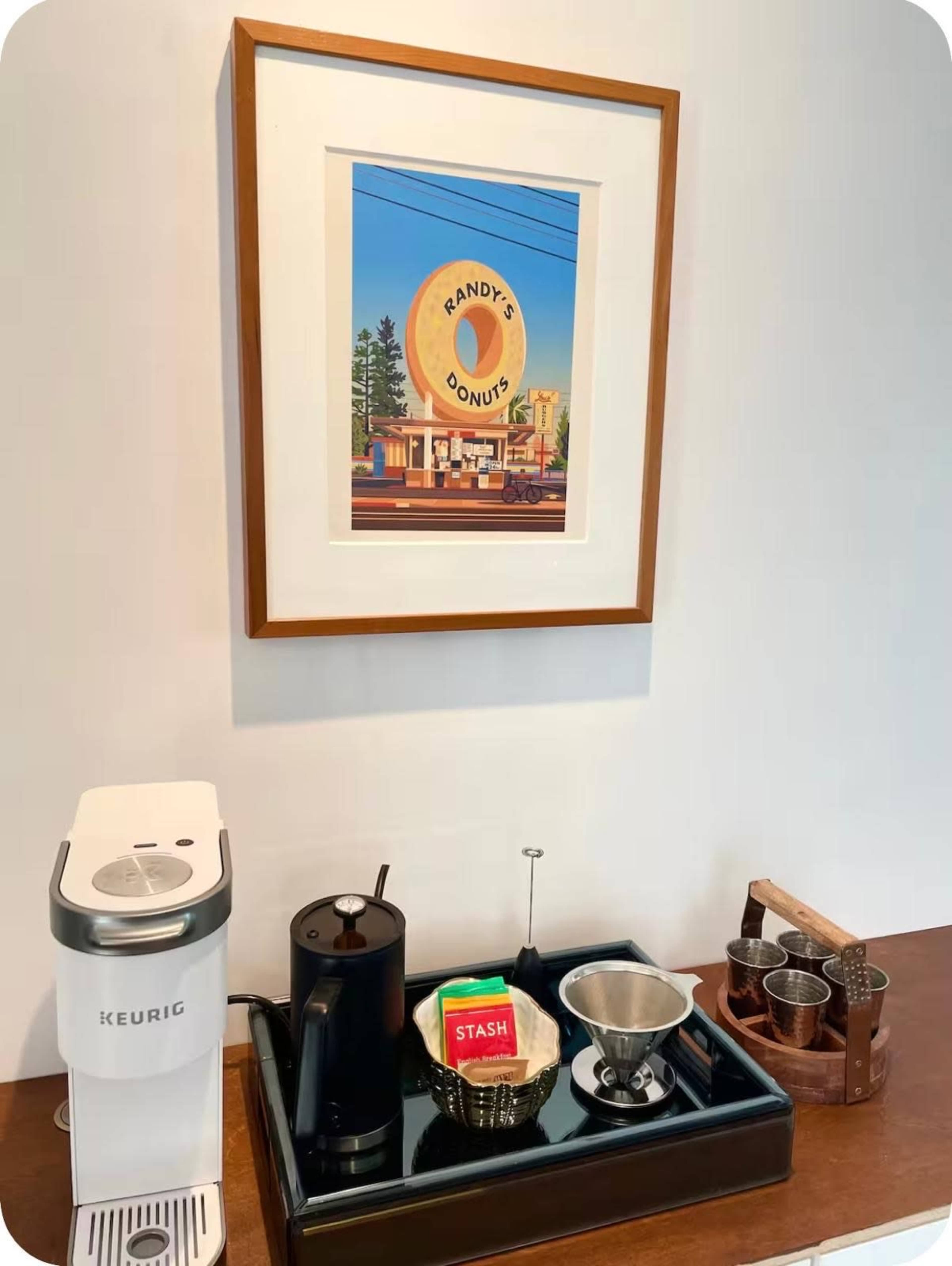 The image shows a coffee station featuring a Keurig machine, a kettle, a tray with tea bags, a filter, and a framed art piece of Randy's Donuts on the wall.