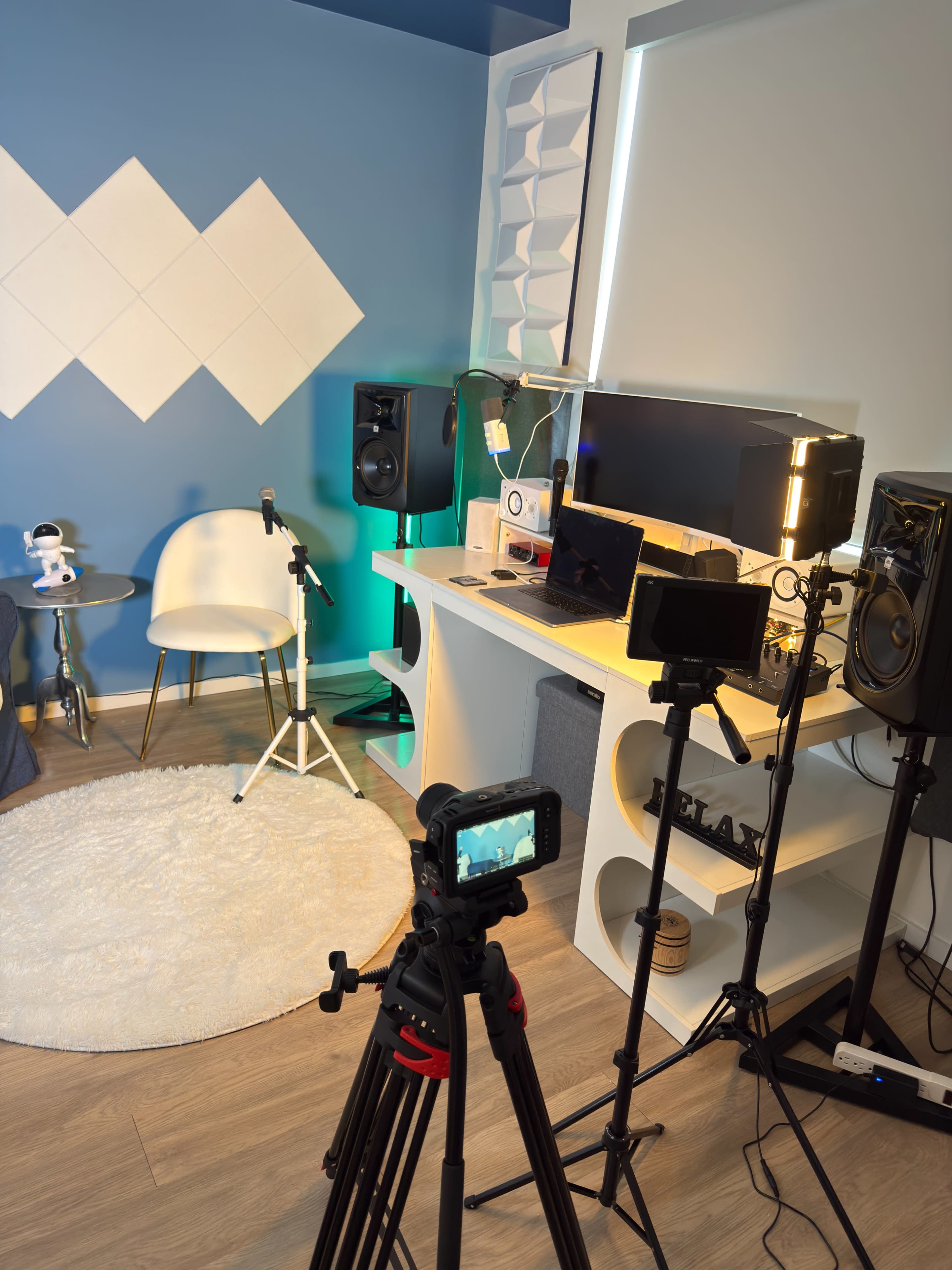 The image shows a modern recording studio setup featuring a microphone on a stand, two studio monitors, a camera on a tripod, and a desk with a computer and lighting equipment, all against a blue wall.