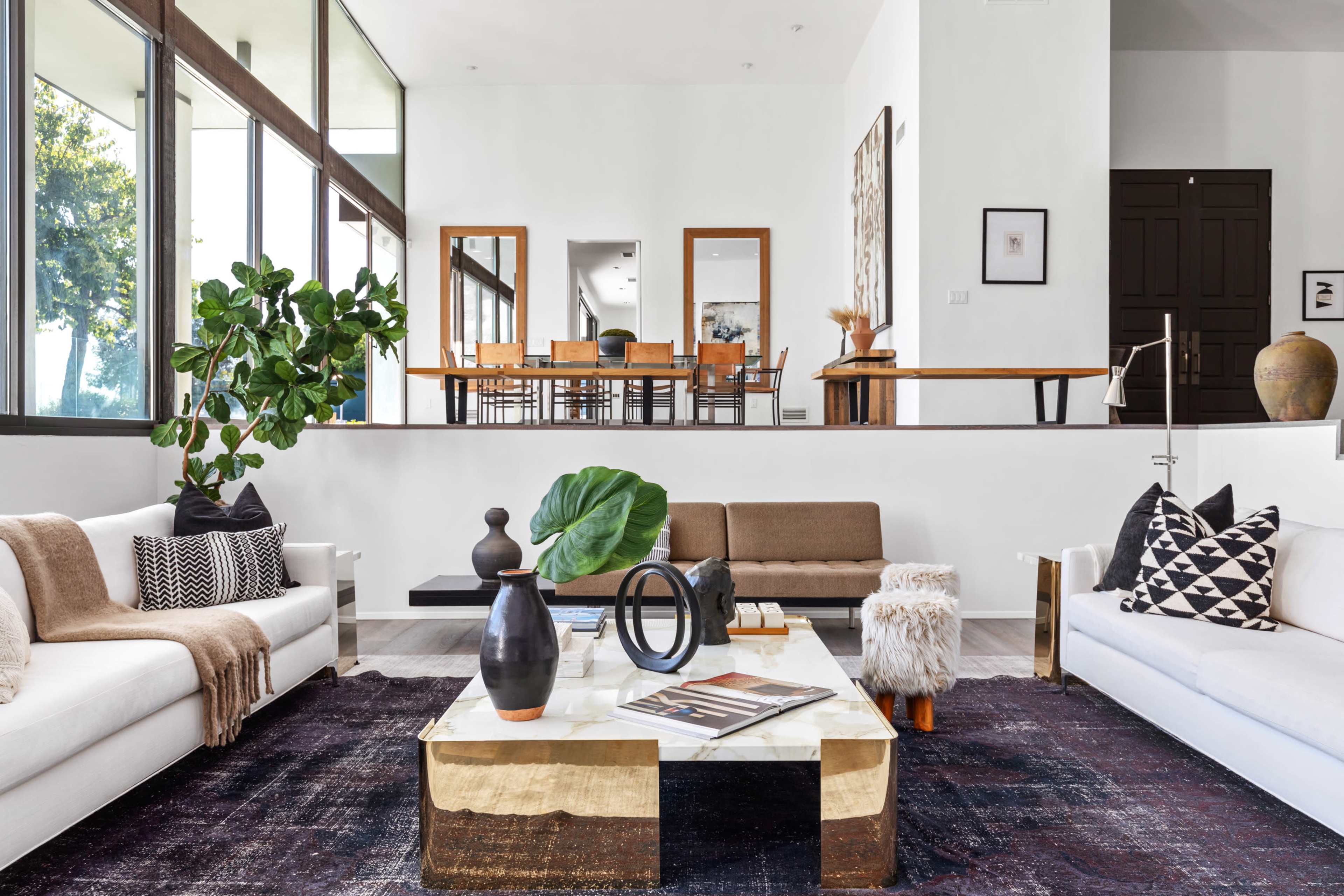 The image shows a modern living room with white sofas, a wooden coffee table, and a view of a dining area through large windows.