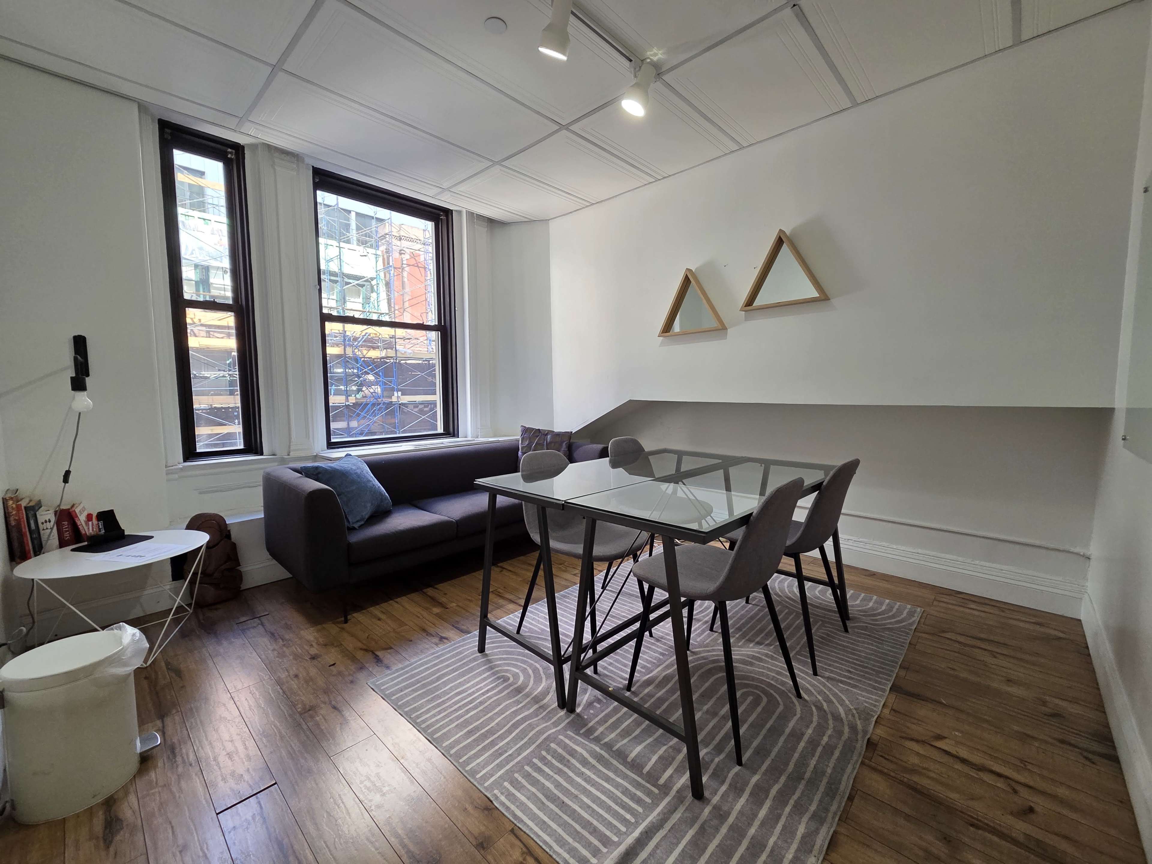 The image shows a modern, minimalist workspace featuring a sofa, a glass dining table with four chairs, and triangular wall decorations, with natural light coming in through a large window.