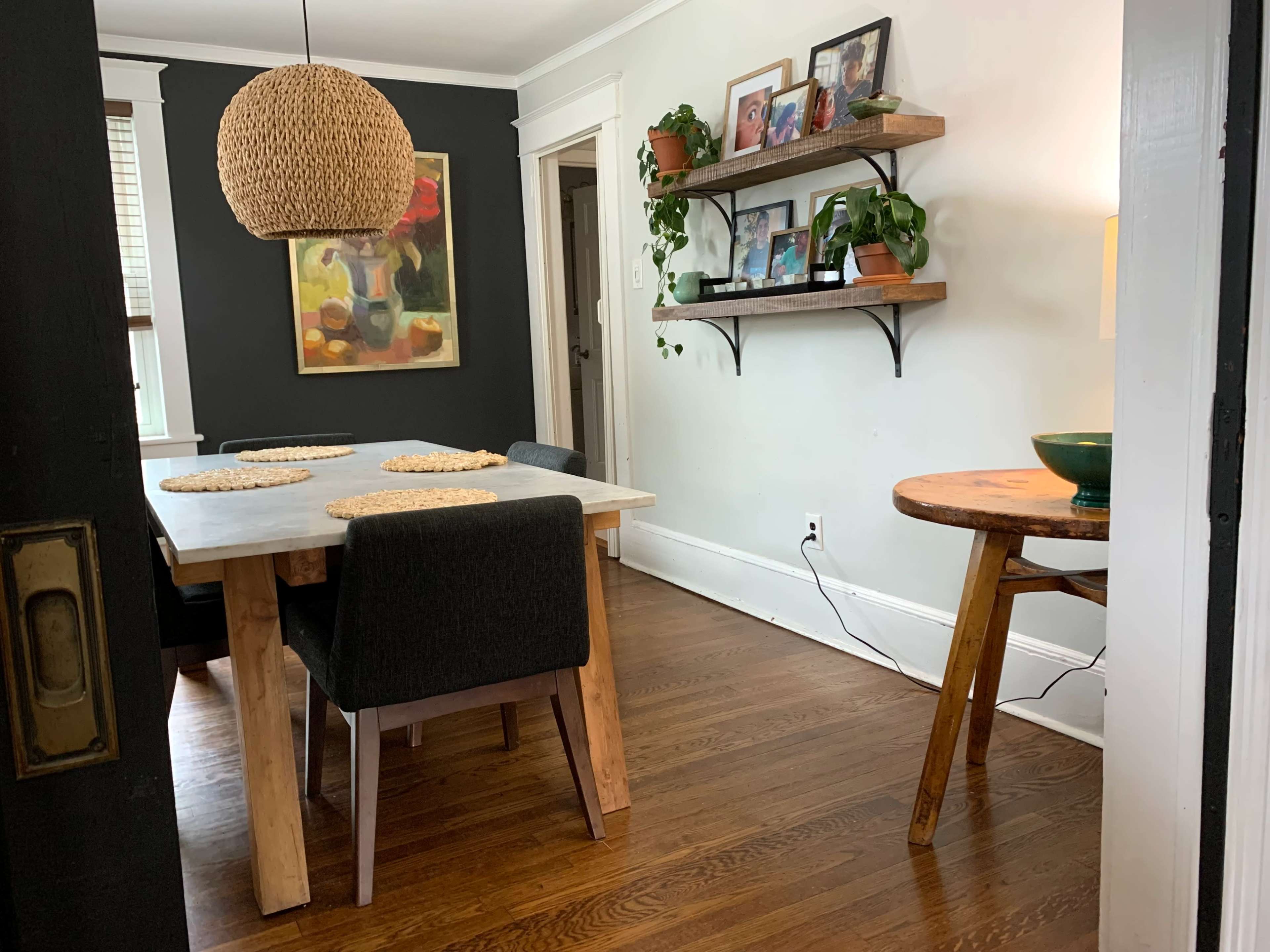A dining area with a wooden table, black chairs, a woven light fixture, and a wall adorned with shelves holding plants and framed pictures.