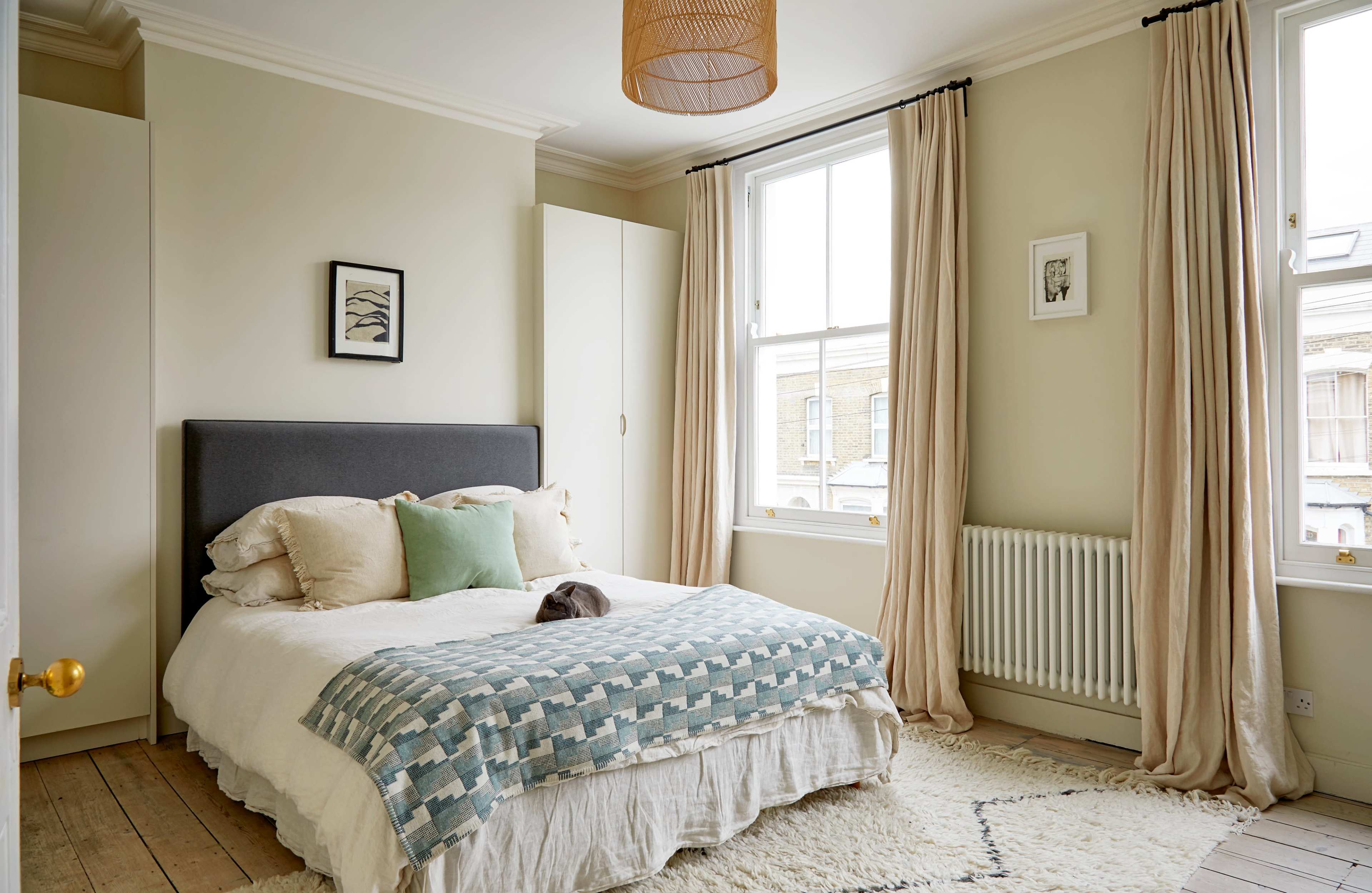 The image shows a neatly arranged bedroom featuring a bed with a light-colored quilt, pillows, and a wooden floor, along with a large window, radiator, and curtains.