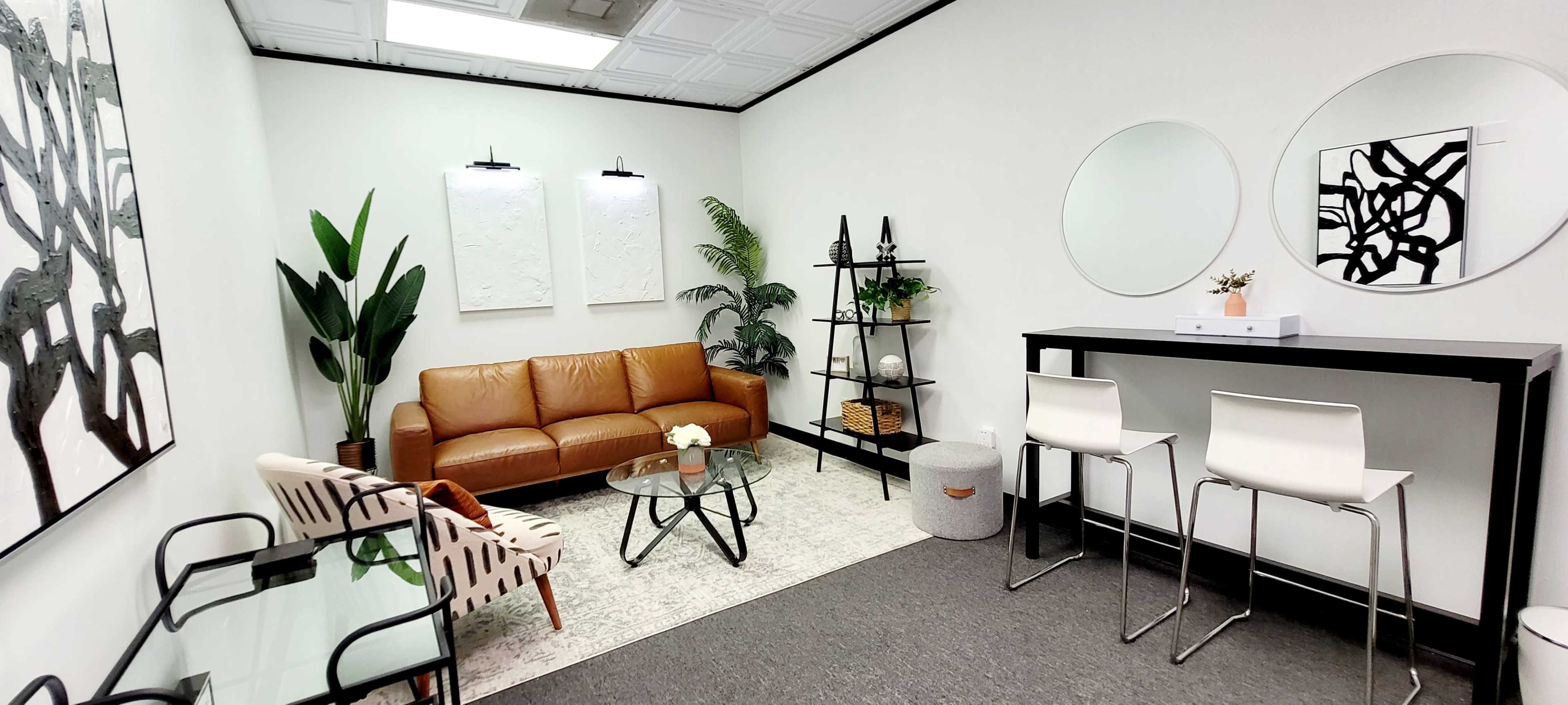 A modern waiting area featuring a brown leather sofa, a glass coffee table, a small round table with a plant, and a bar-style seating area with white chairs.