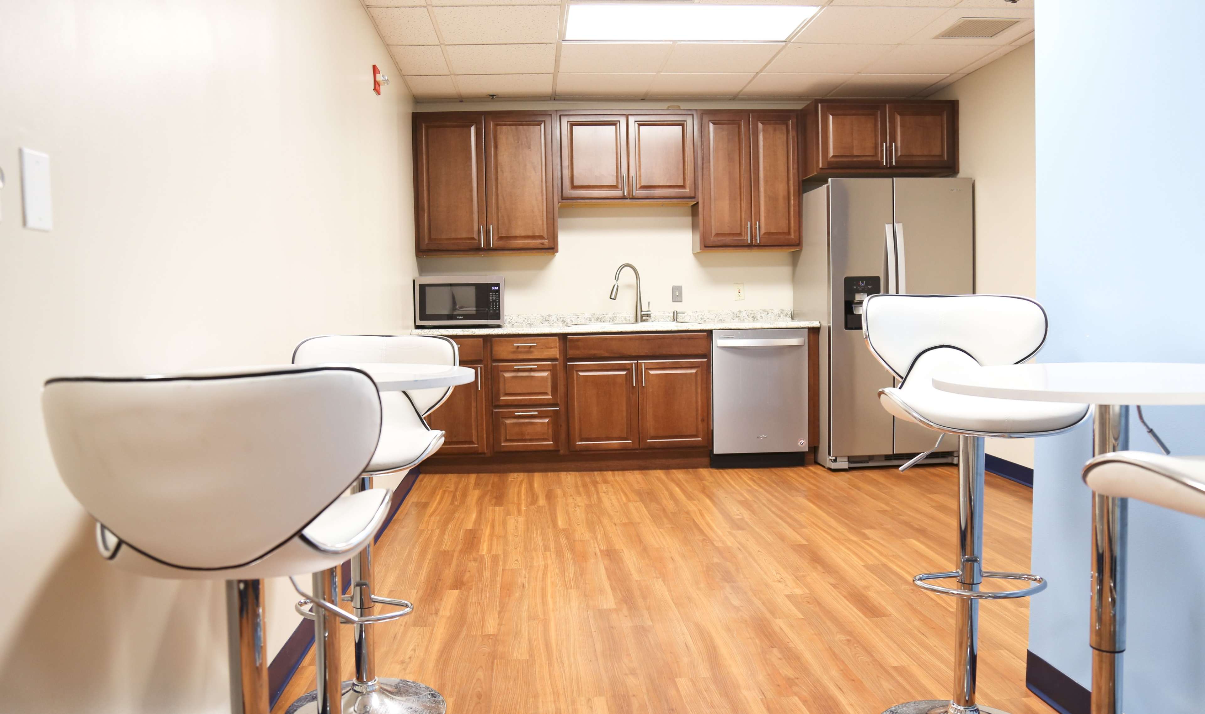 A modern kitchen features wooden cabinets, stainless steel appliances, and white barstools around a small table.