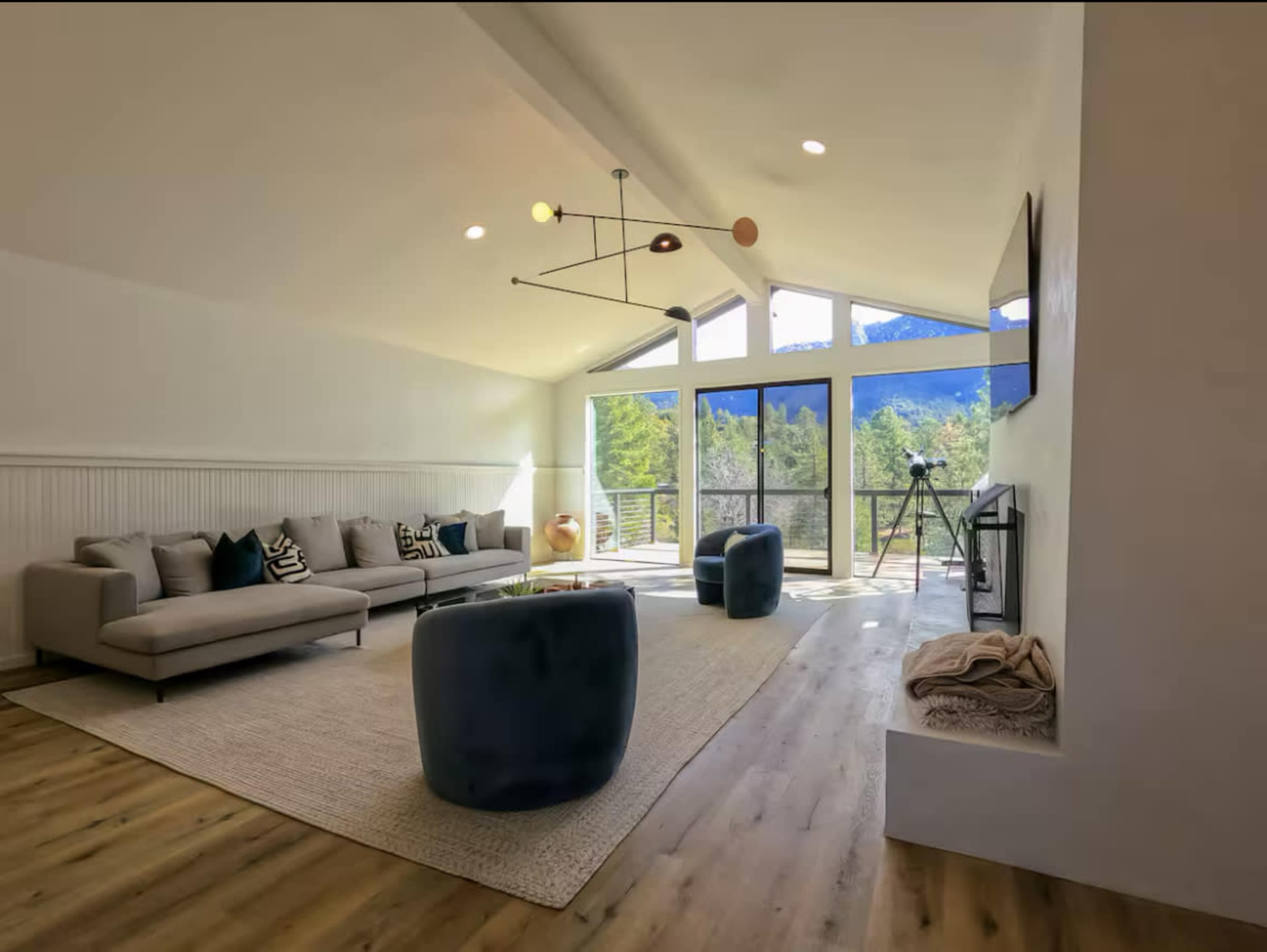 The image shows a modern living room with a light-colored sofa, two dark blue ottomans, and large windows that overlook a mountain view.