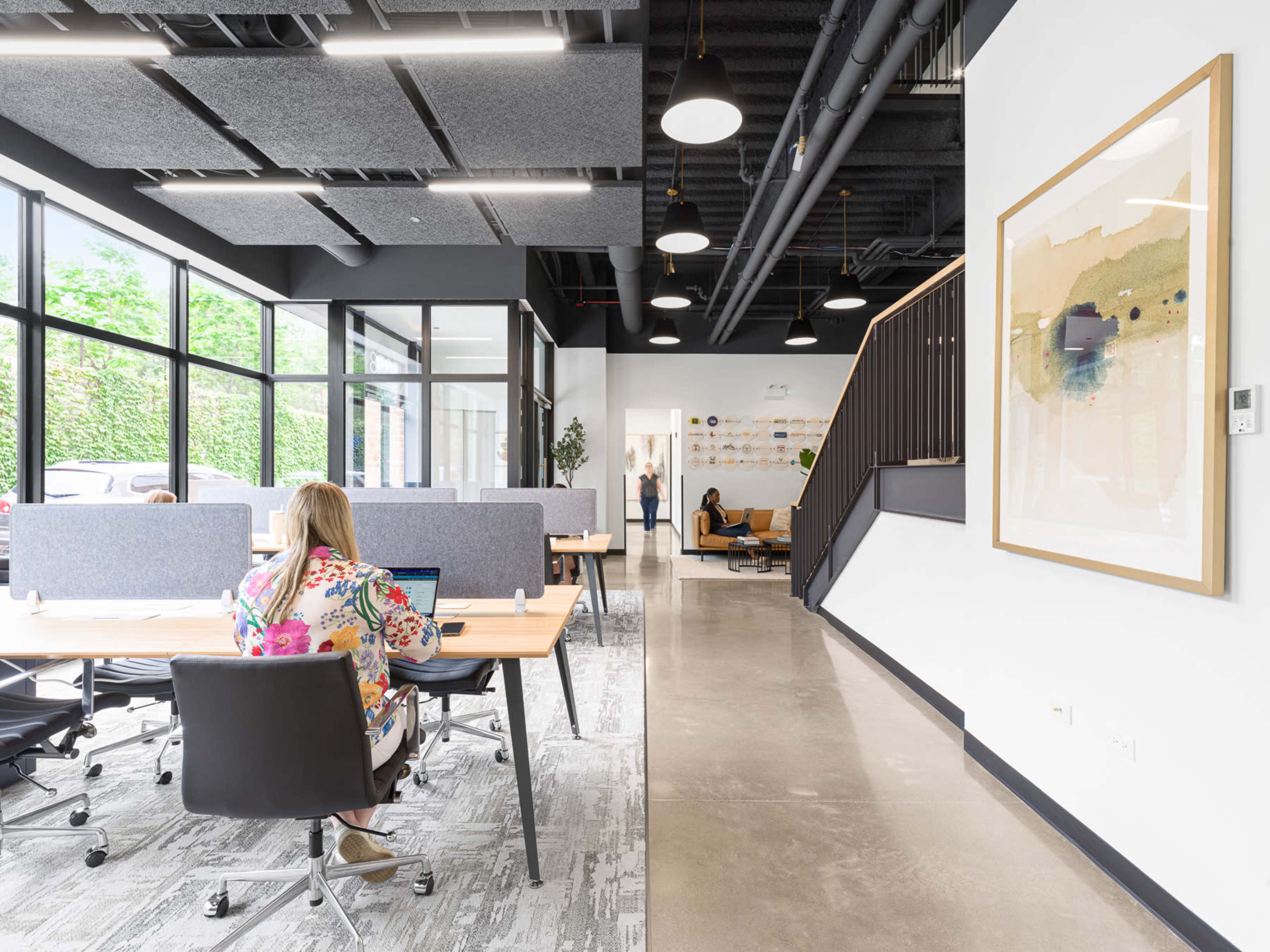 A modern open office space features desks with dividing panels, a person working on a laptop, and a staircase leading to a lounge area.
