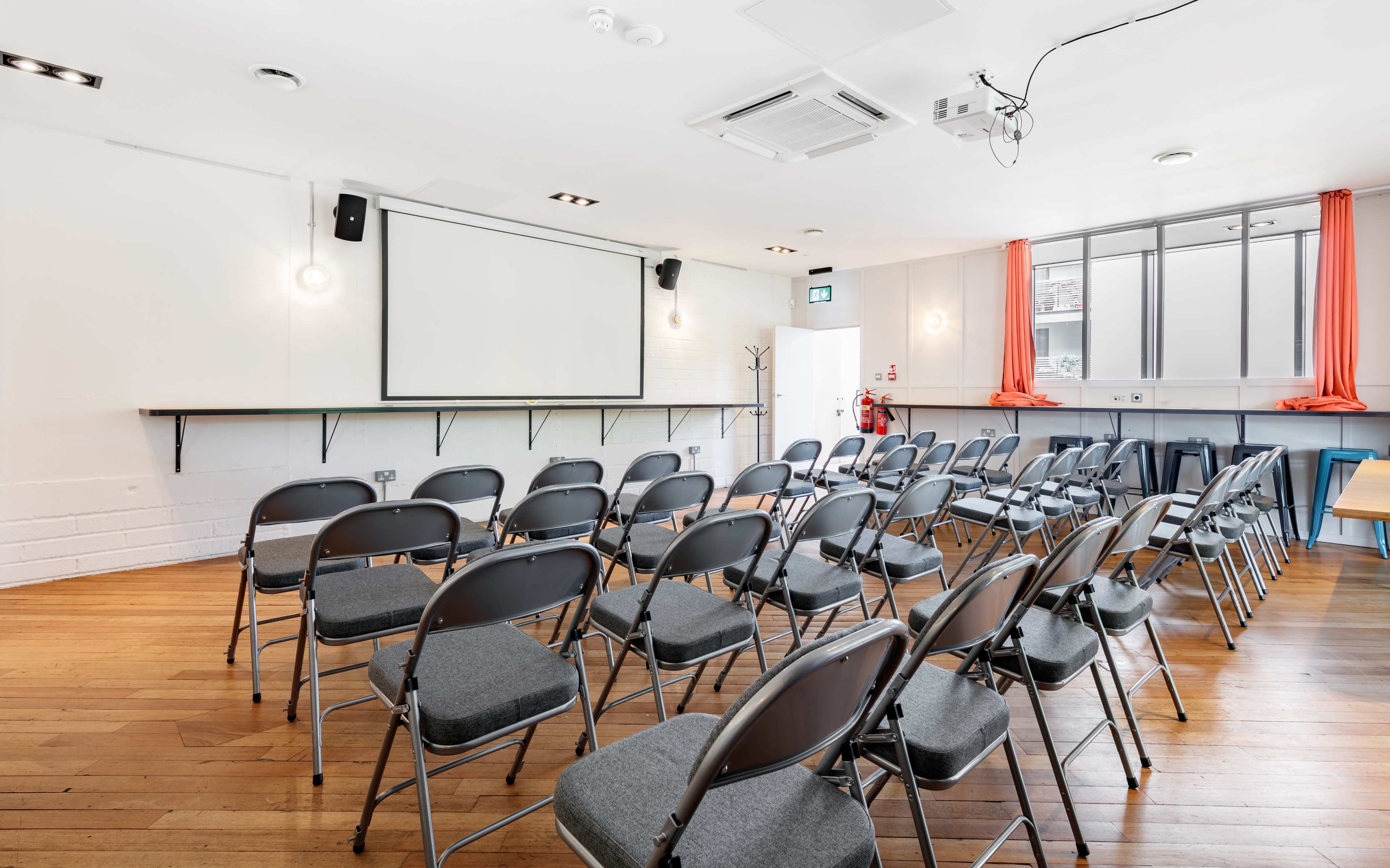 A meeting room features rows of metal chairs facing a projection screen, with large windows draped in orange curtains and polished wooden flooring.