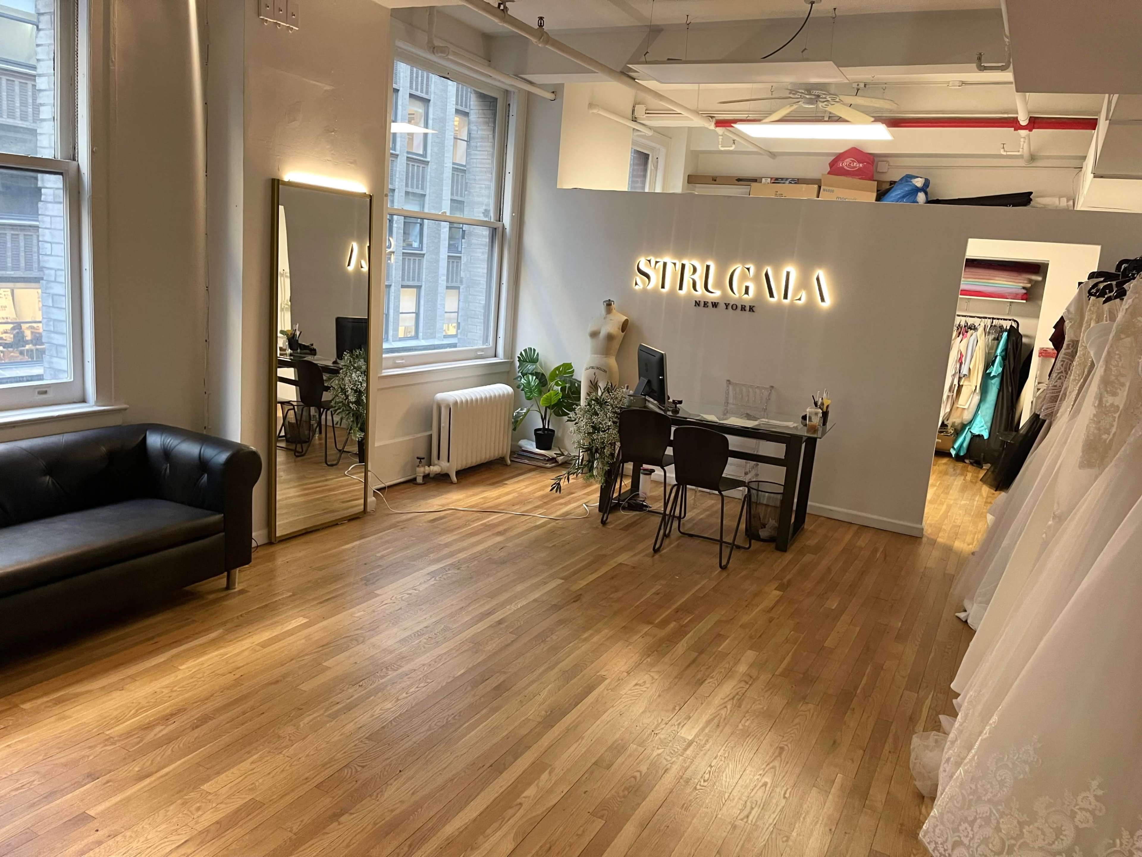 The image shows a spacious studio with a black leather sofa, a desk with a plant, a large mirror, and a clothing rack displaying various dresses.