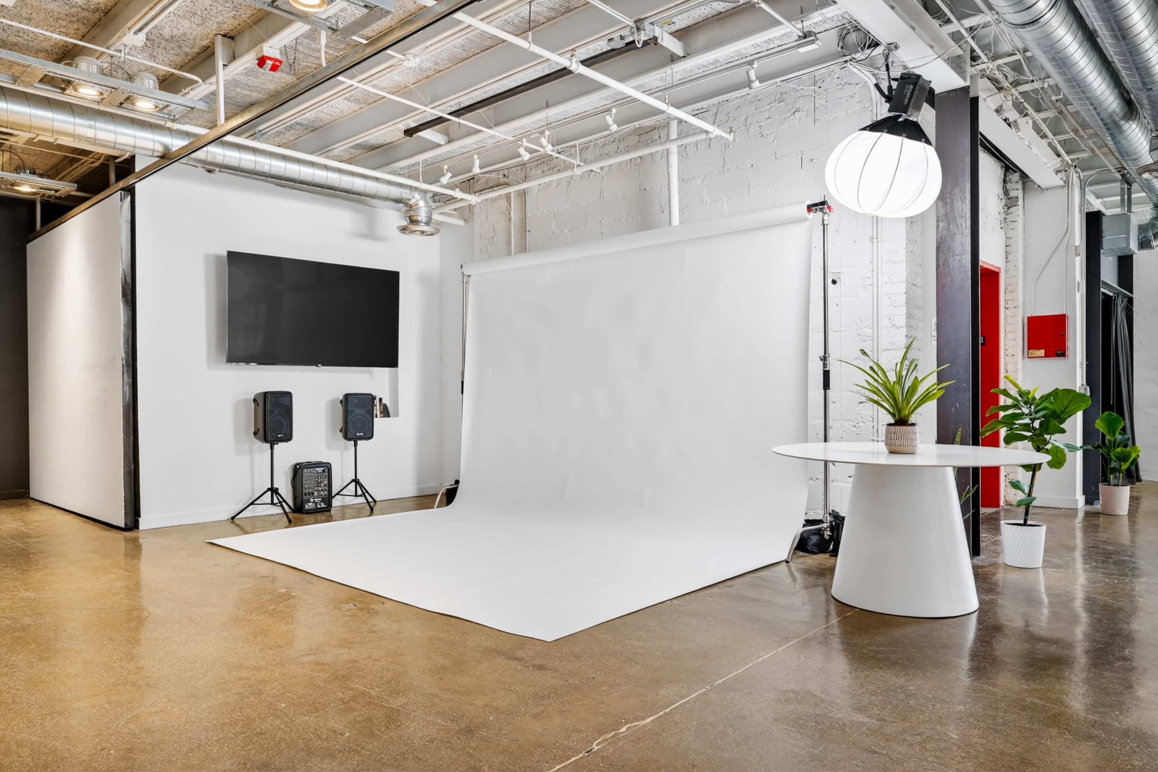 The image shows a studio space featuring a white backdrop for photography, a wall-mounted TV, two speakers, and a round table with a plant on it.