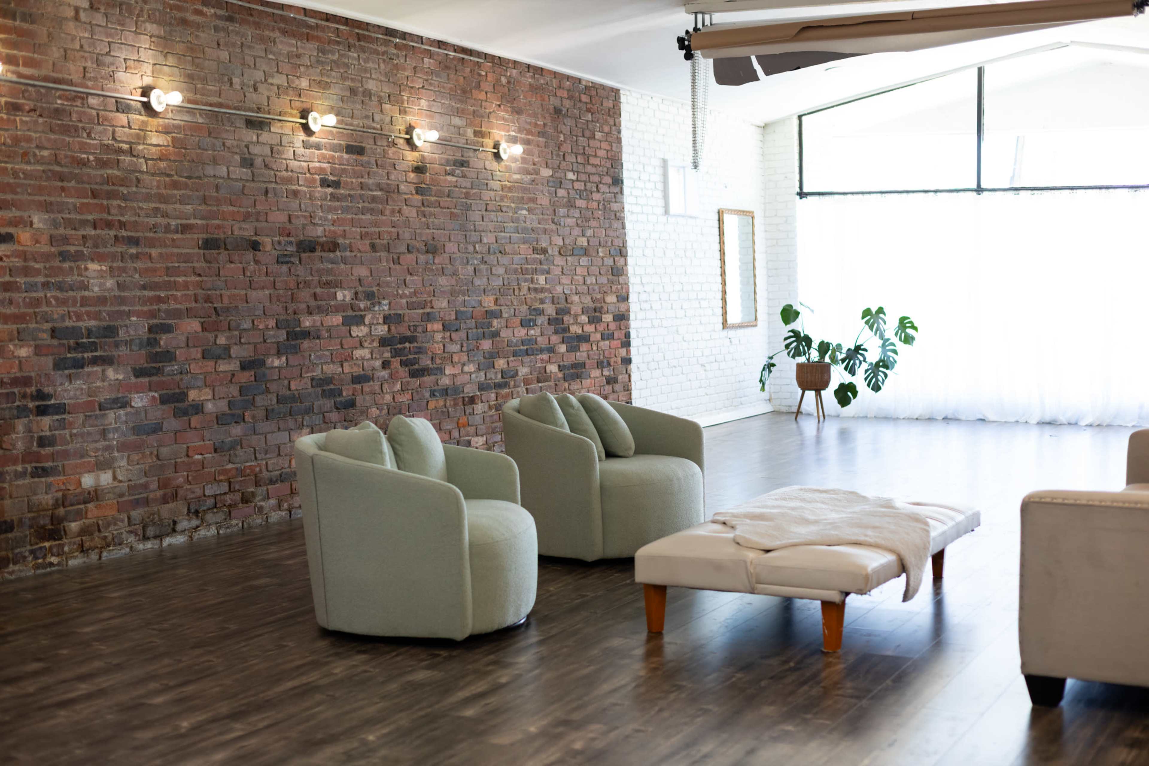 The image shows a spacious room with exposed brick walls, two light green armchairs, a low table with a soft cover, and a plant in the corner.