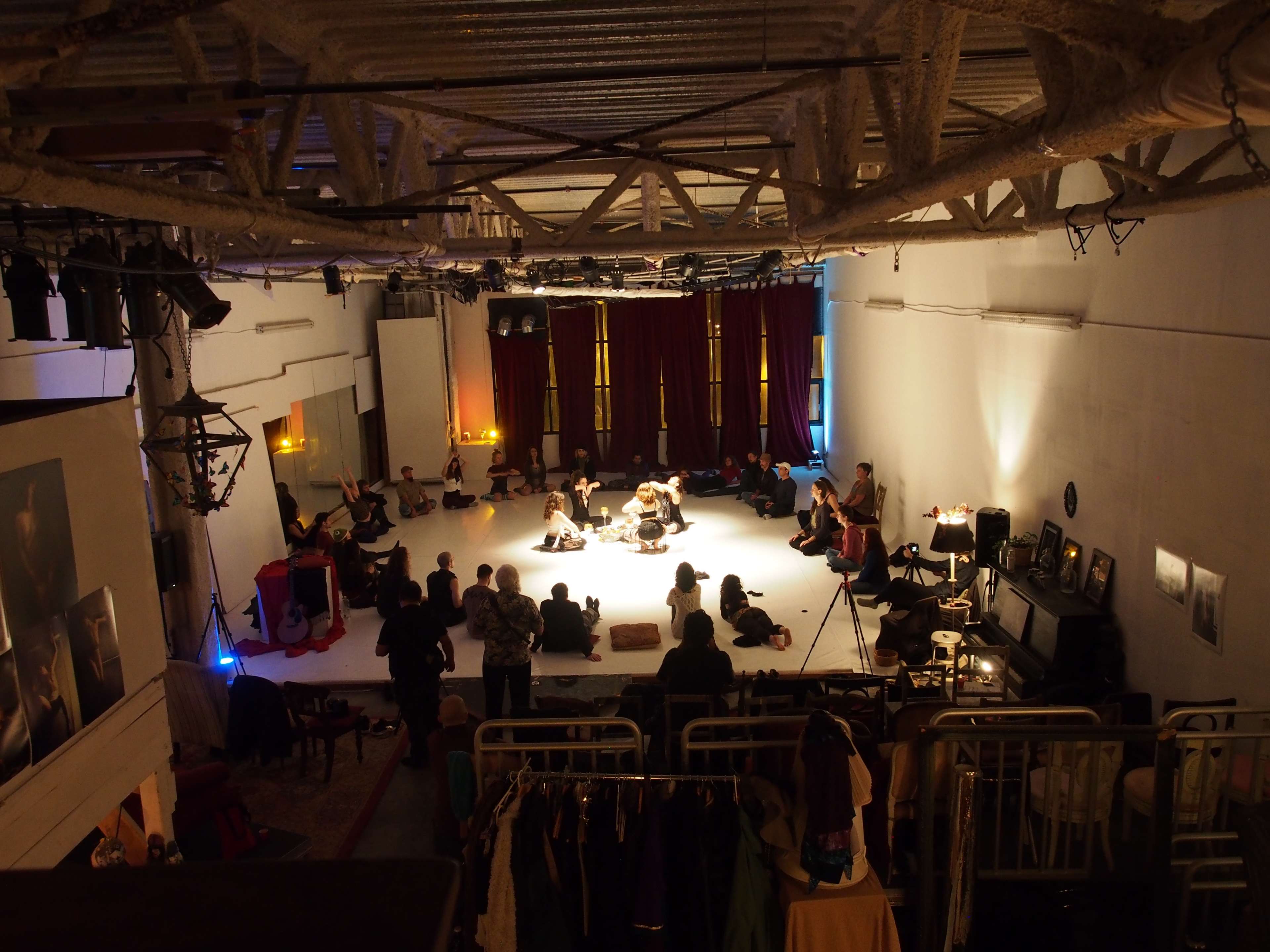 A large group of people is seated around a performance space in a dimly lit room with raised ceilings and red curtains.