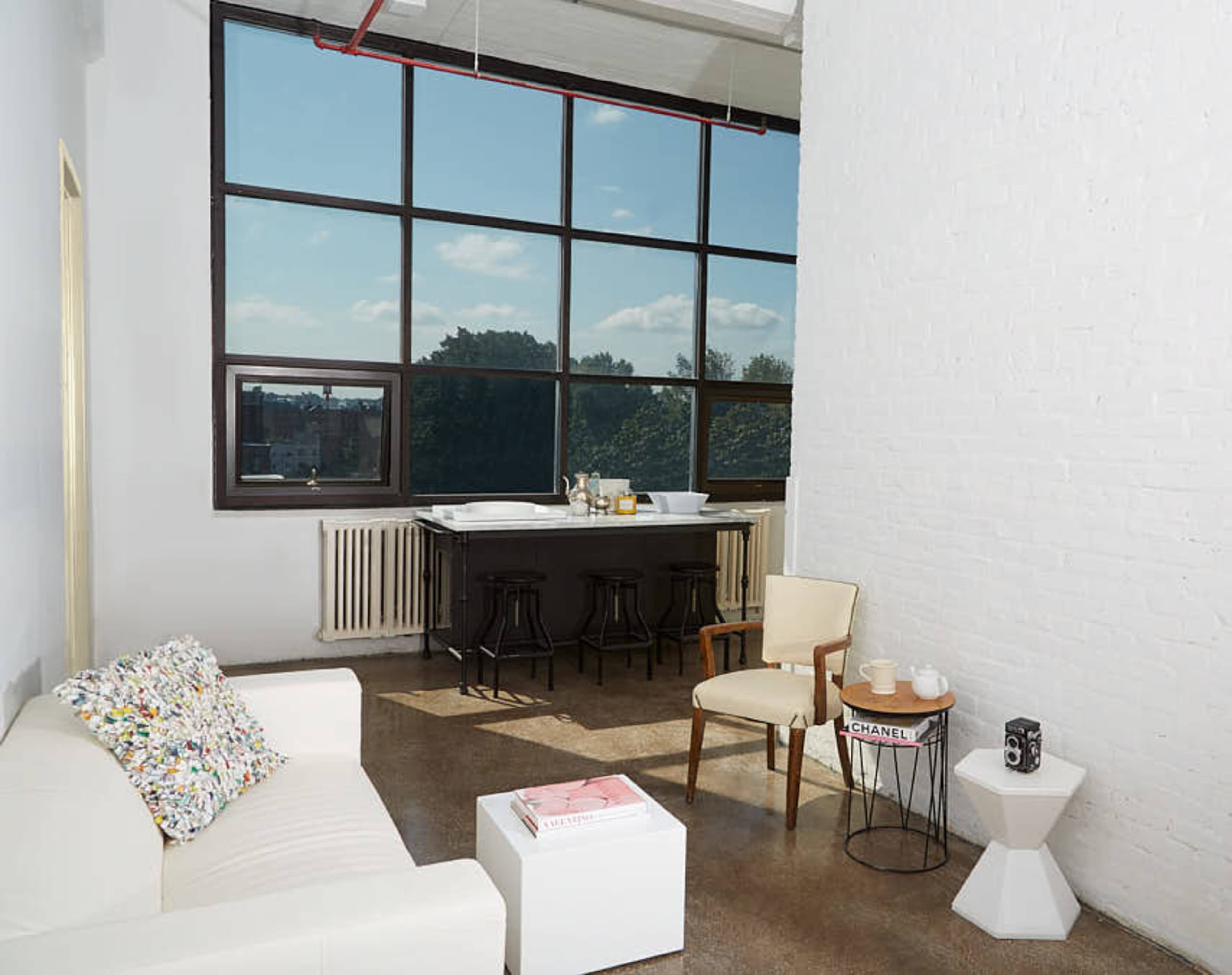 A bright room features a white sofa, a small table, two chairs, and a large window overlooking trees.
