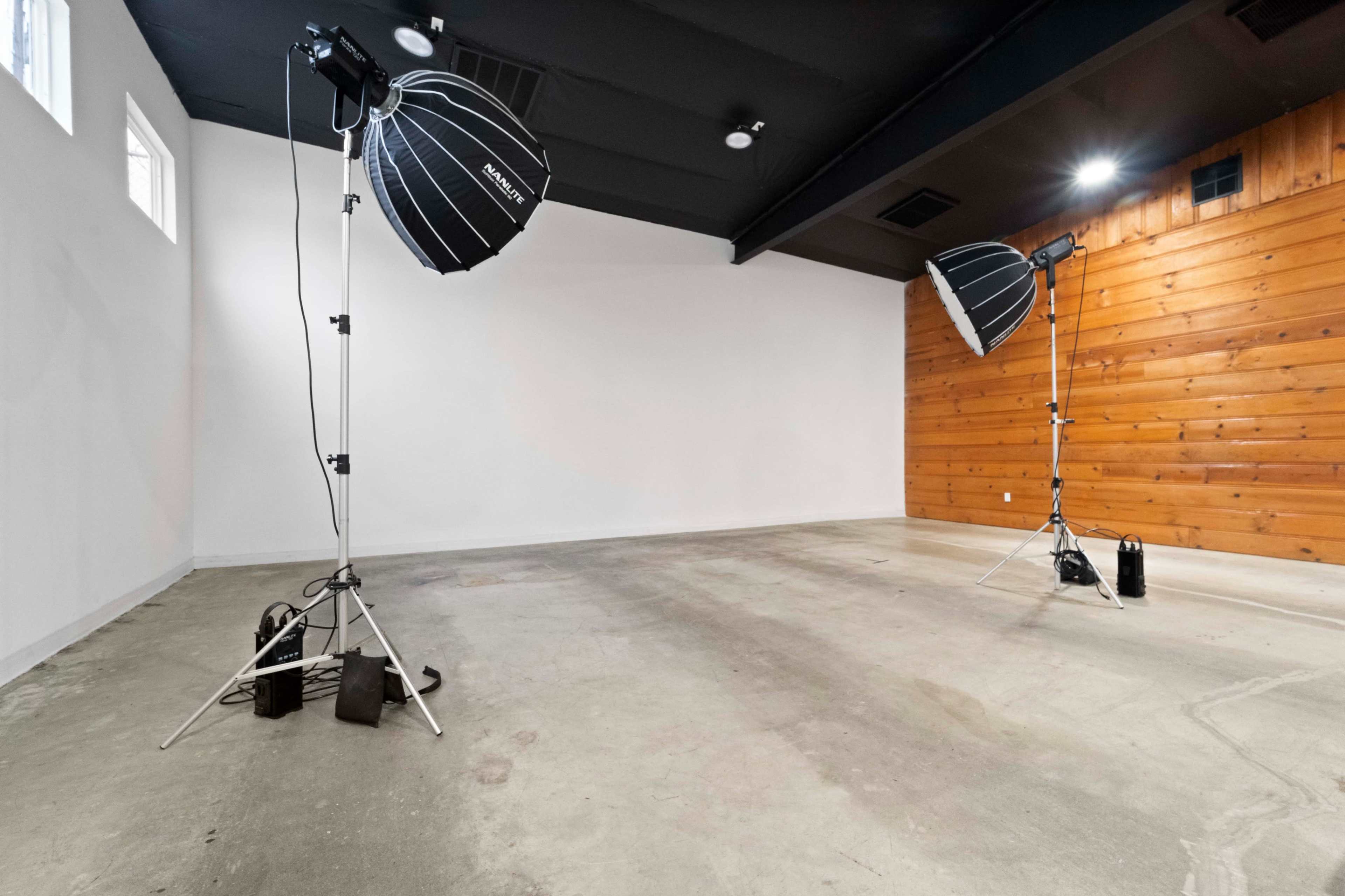 A minimalist studio space with two large light modifiers set up on stands, against a backdrop of white walls and a wooden accent wall.