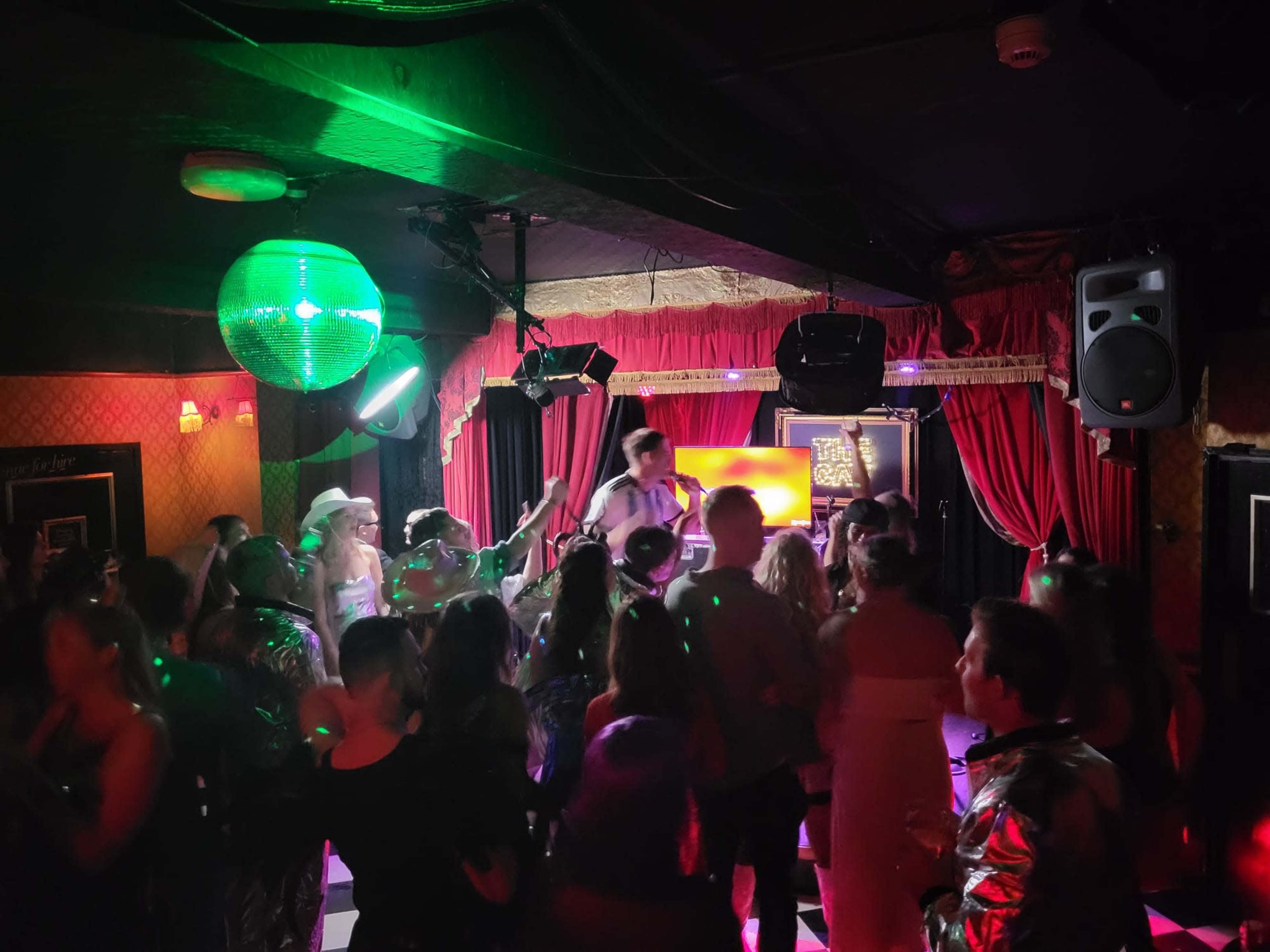 A lively crowd dances under a disco ball in a dimly lit bar with colorful lights.