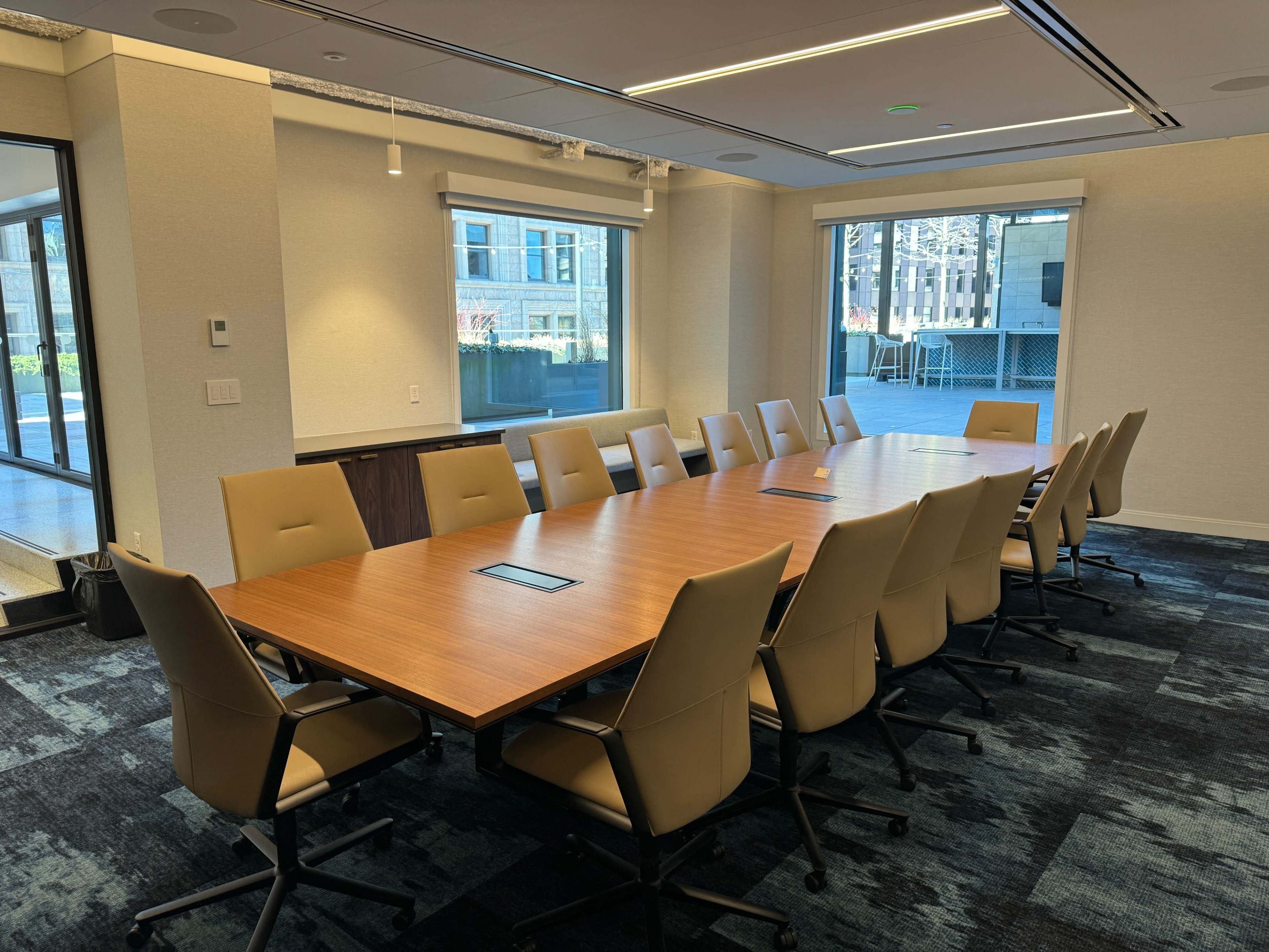 A spacious conference room features a long wooden table surrounded by twelve beige chairs, with large windows providing natural light.