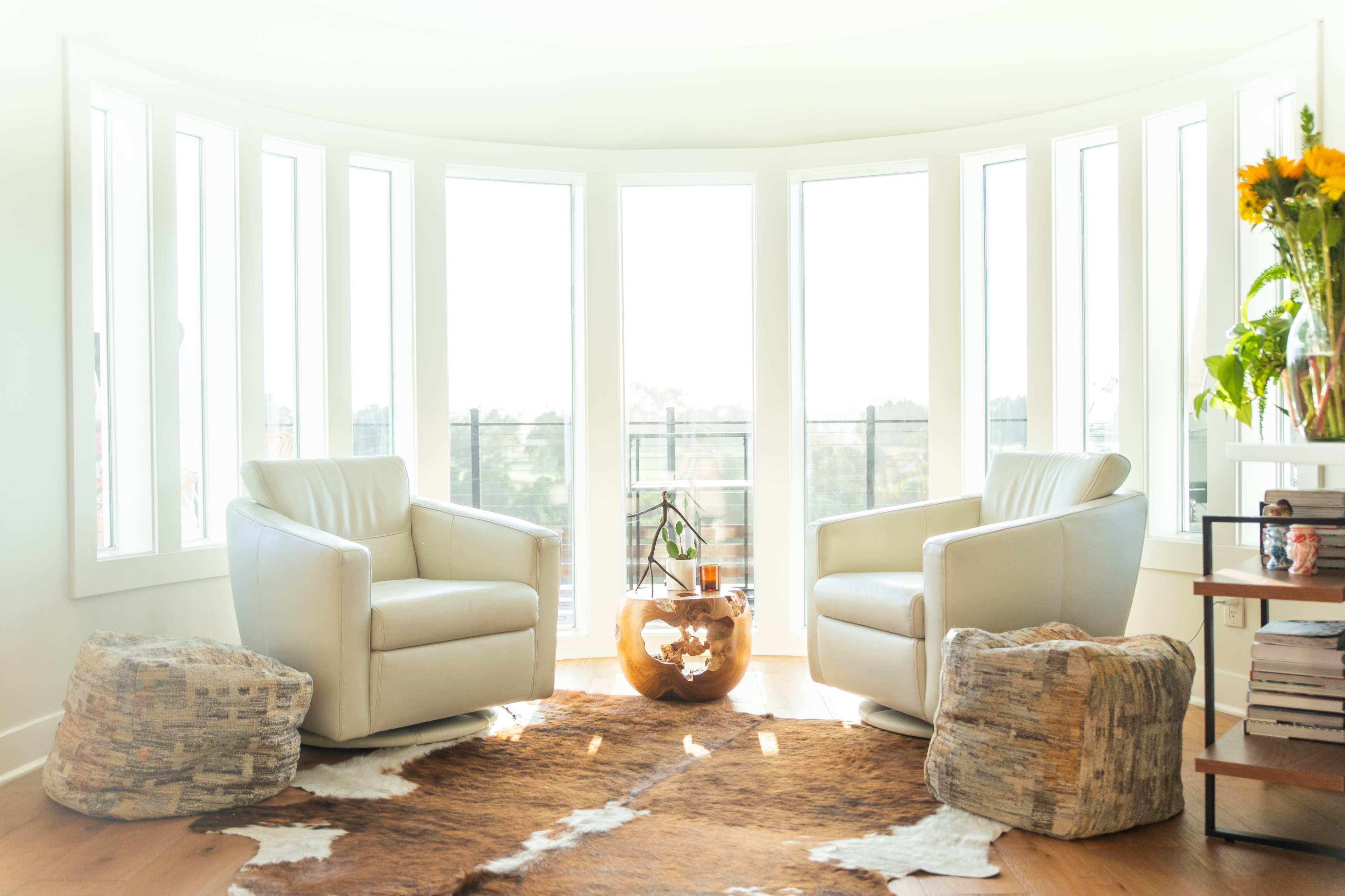 A bright room with two white armchairs facing each other, surrounded by large windows and a round coffee table, with a rug made of animal hide on the wooden floor.
