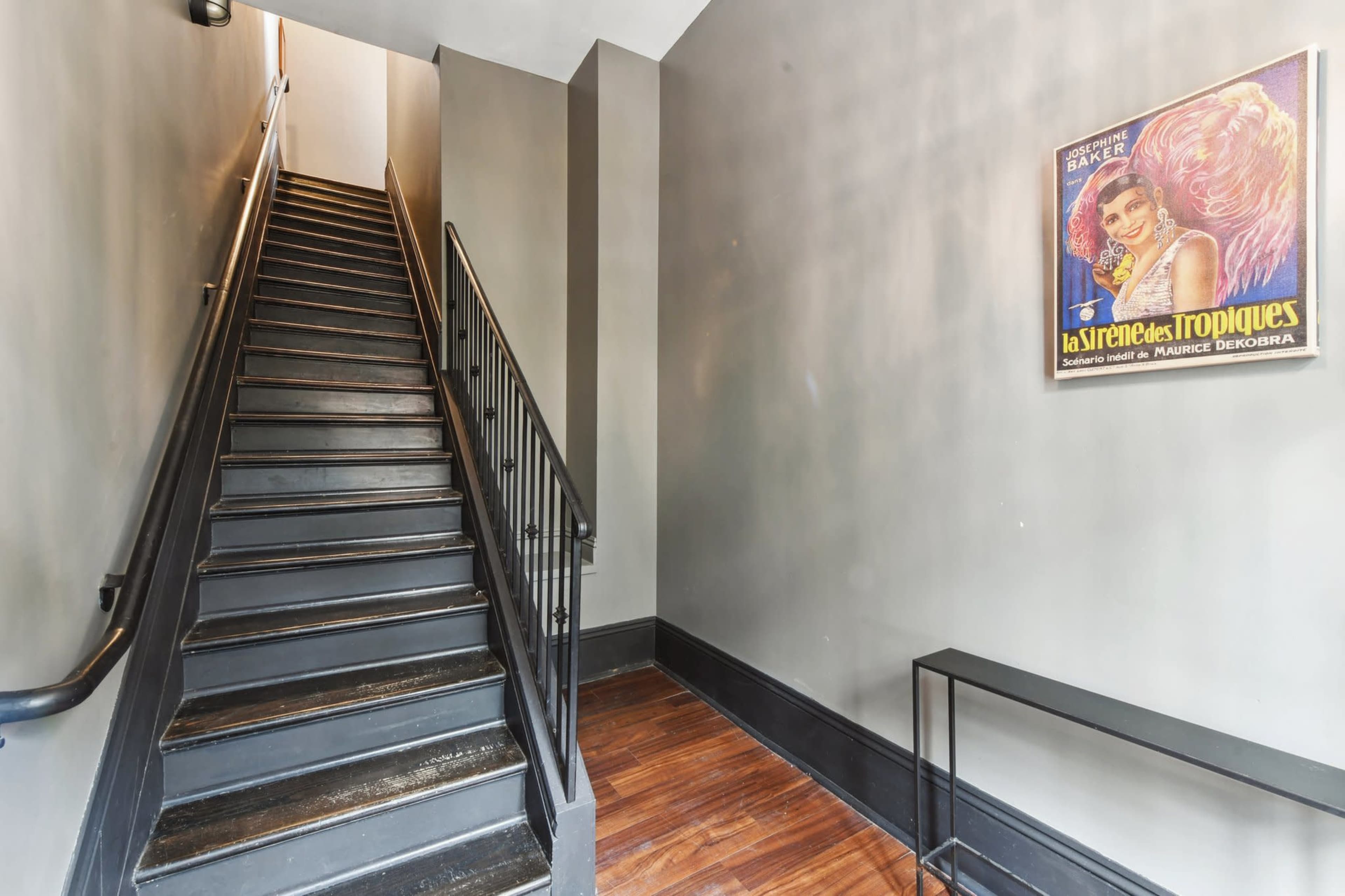 The image shows a staircase with black steps leading upward, paired with a framed vintage poster on the wall beside it.