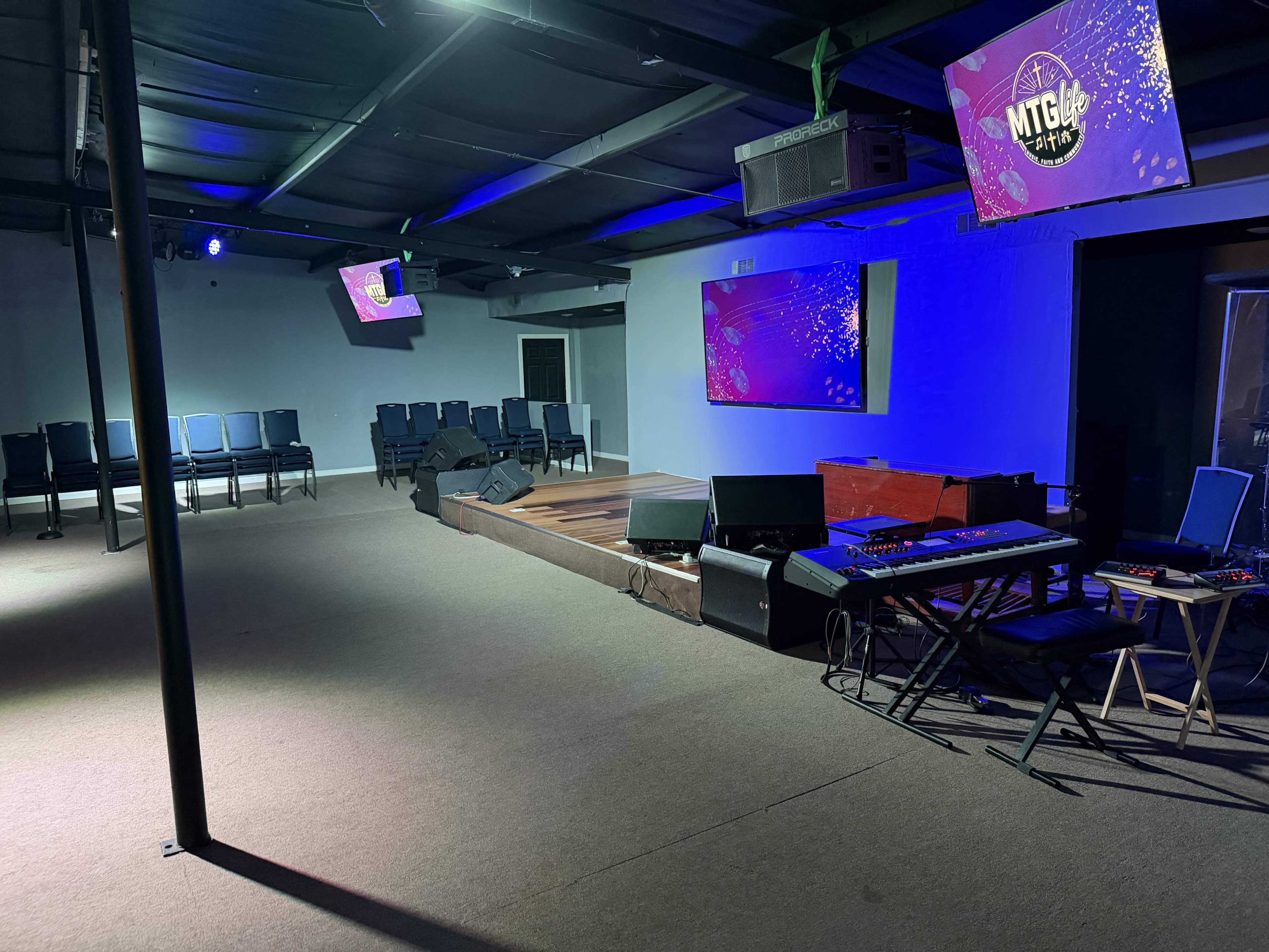 The image shows a dimly lit indoor venue with a performance stage, musical equipment, seating arranged in rows, and two screens displaying graphics on the walls.
