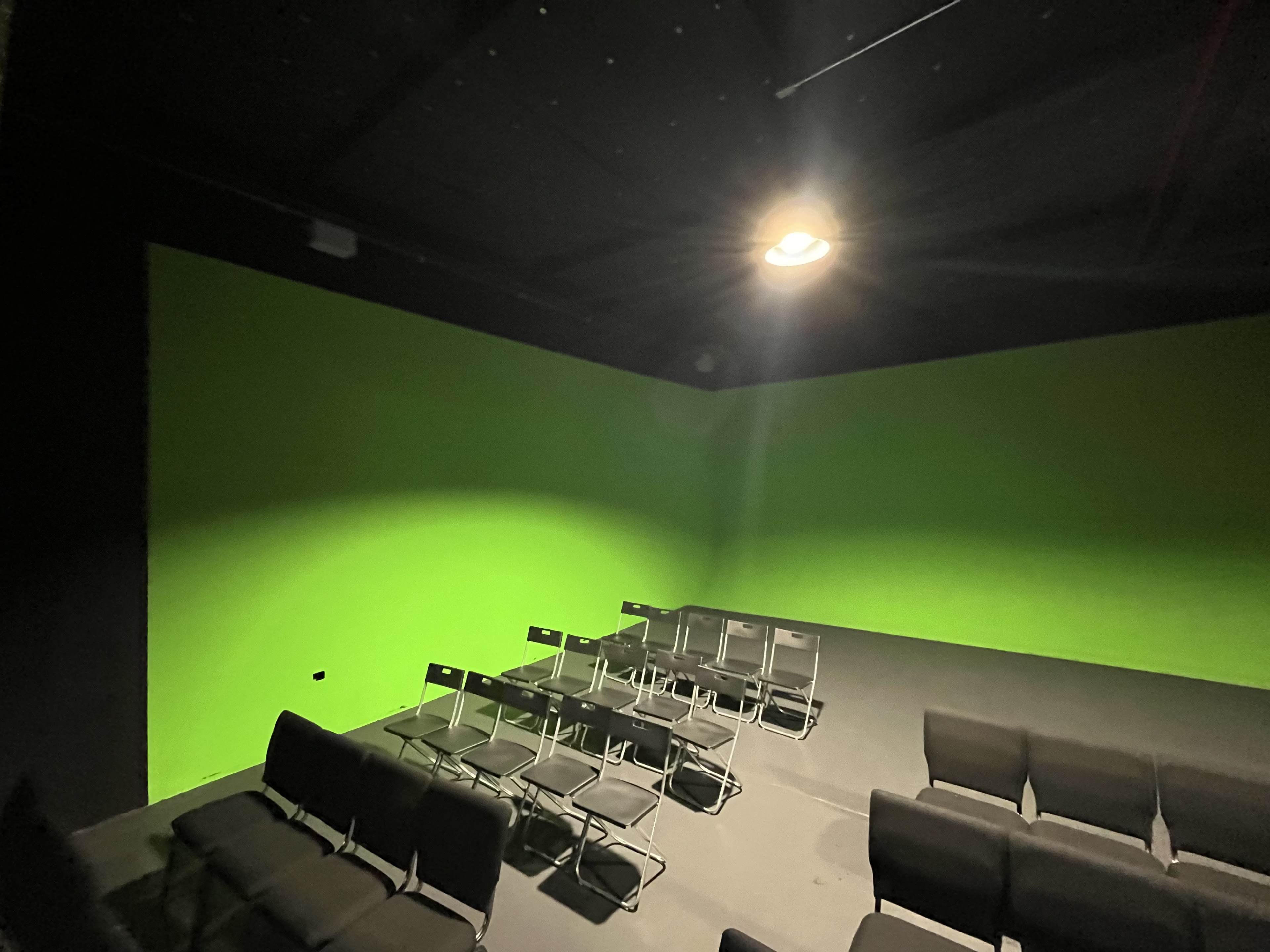 A dimly lit room with black seating arranged in rows facing a bright green wall.
