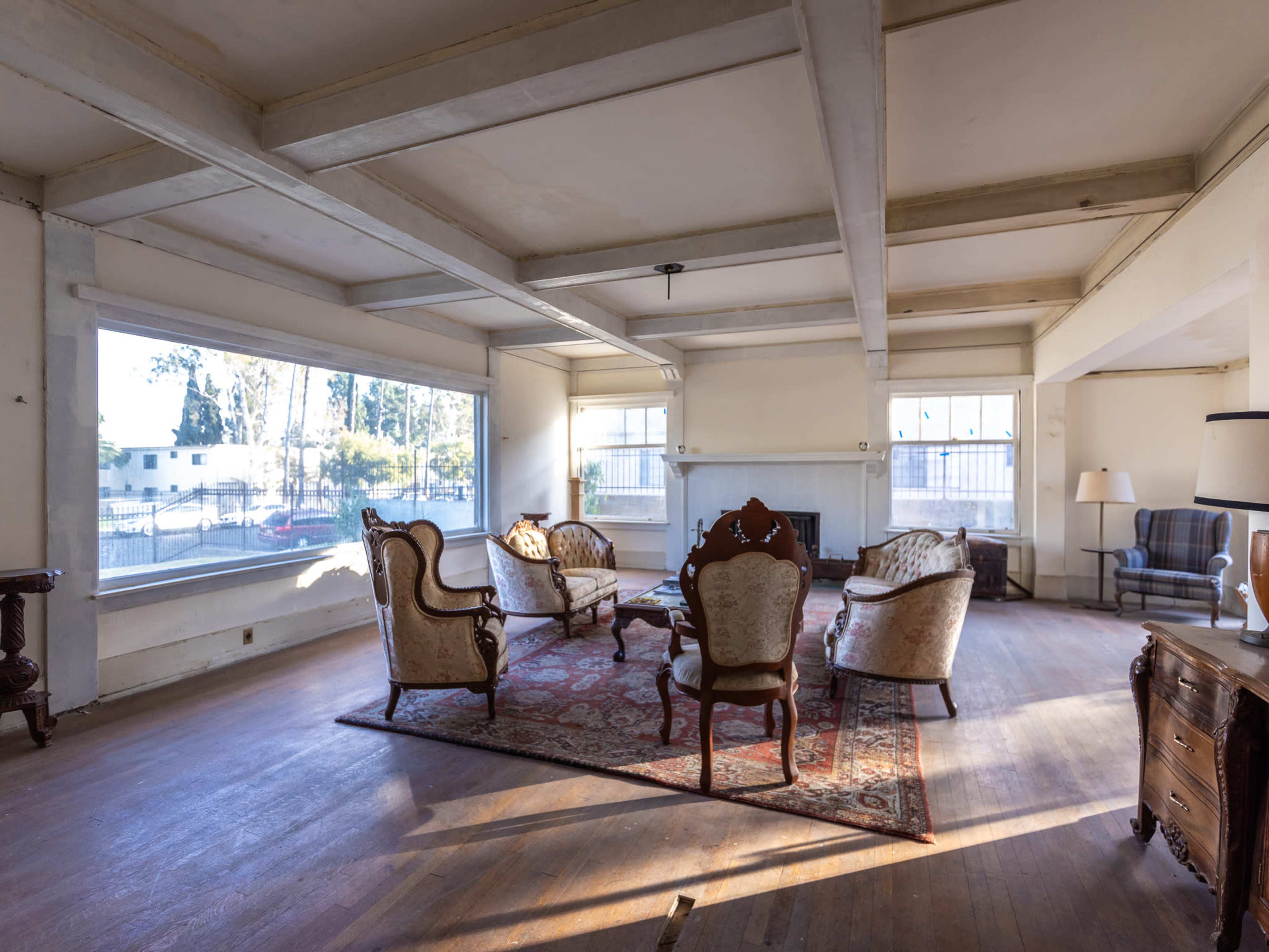 A spacious living room with four upholstered armchairs arranged around a wooden table on a patterned rug, illuminated by natural light from large windows.