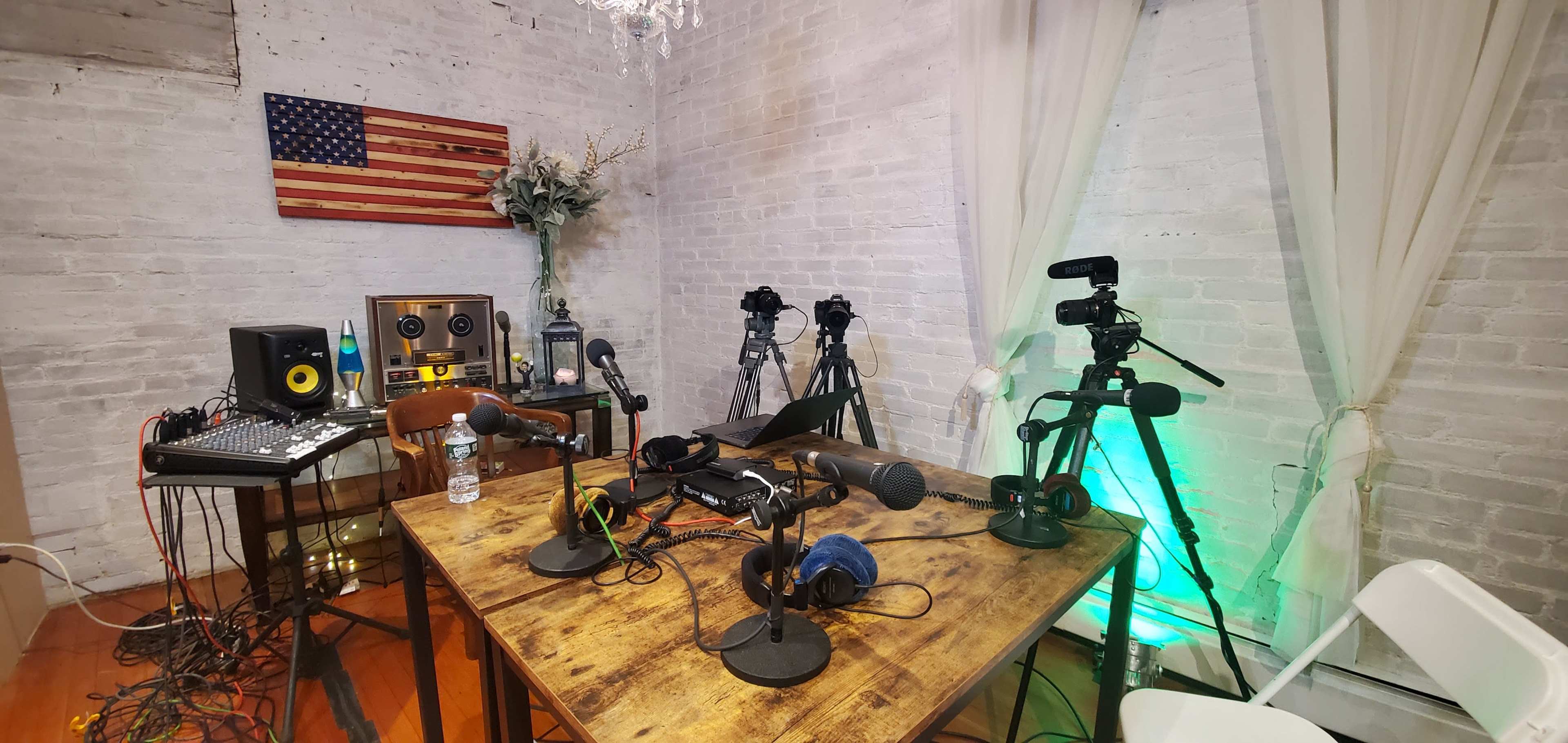 A recording studio setup featuring multiple microphones, cameras, and audio equipment against a backdrop of a wooden flag and a potted plant.