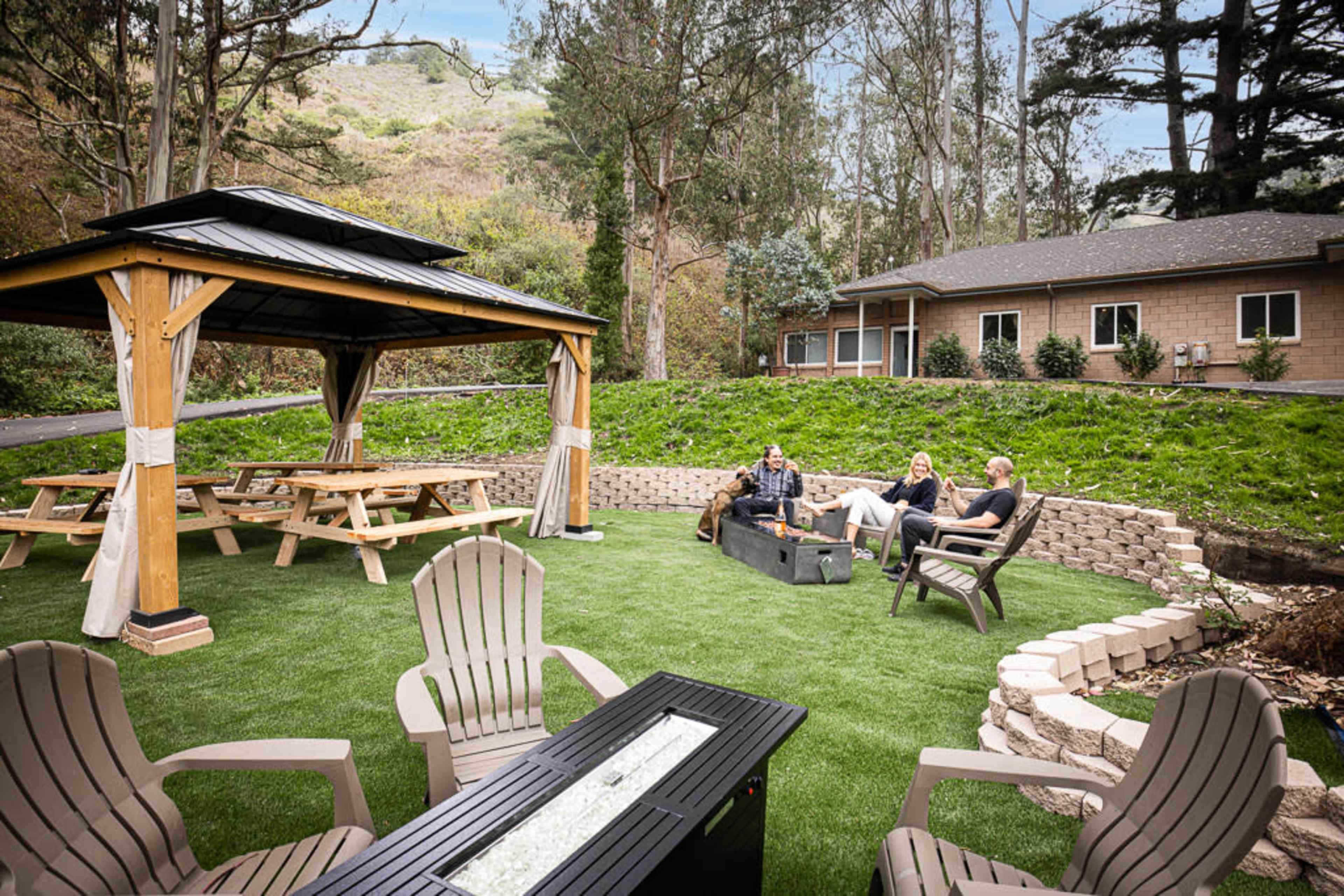 A group of three people sits around a fire pit on a landscaped lawn near a gazebo and a building surrounded by trees.