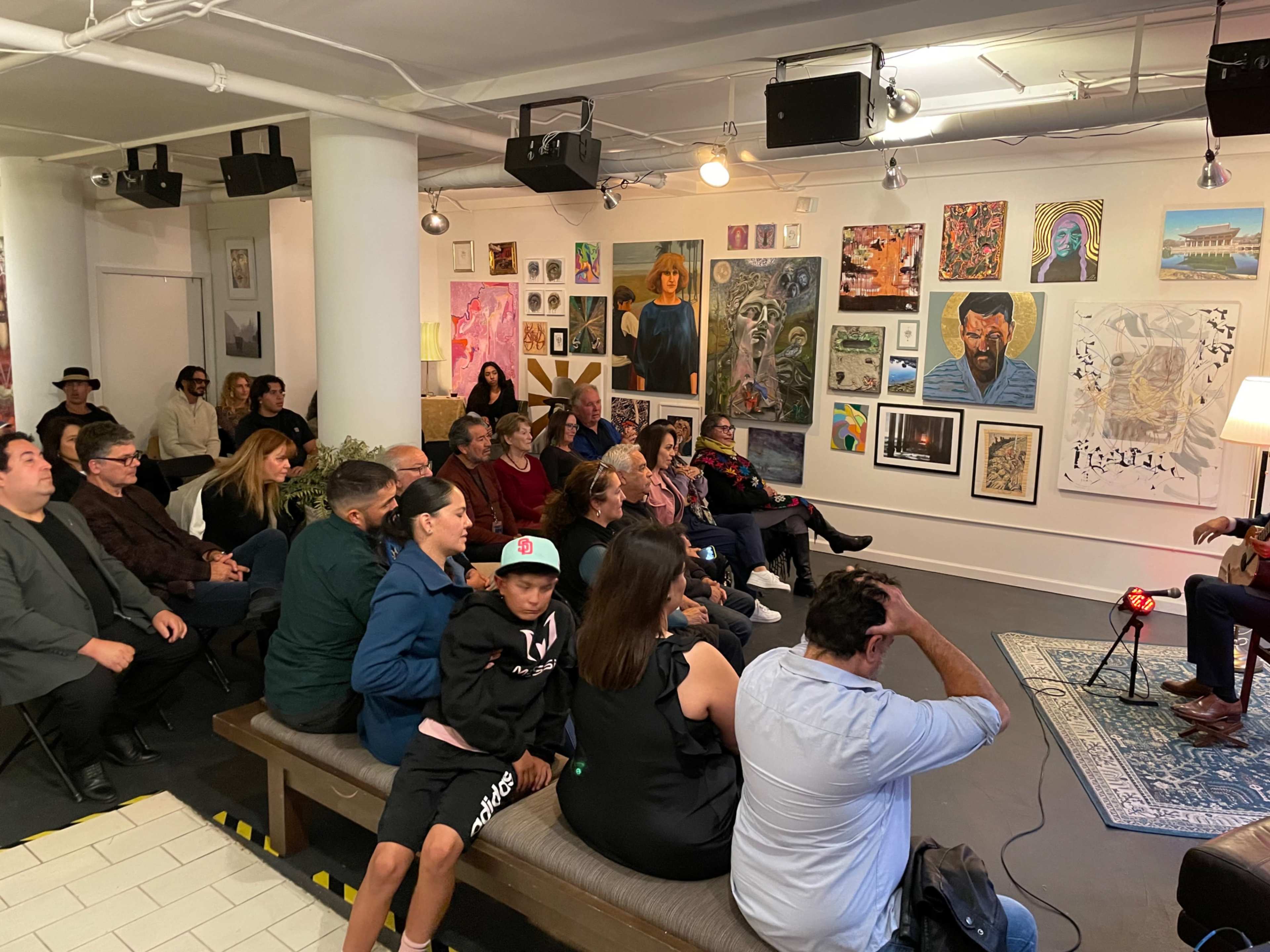 A seated audience listens attentively to a performer in a gallery adorned with various artworks on the walls.
