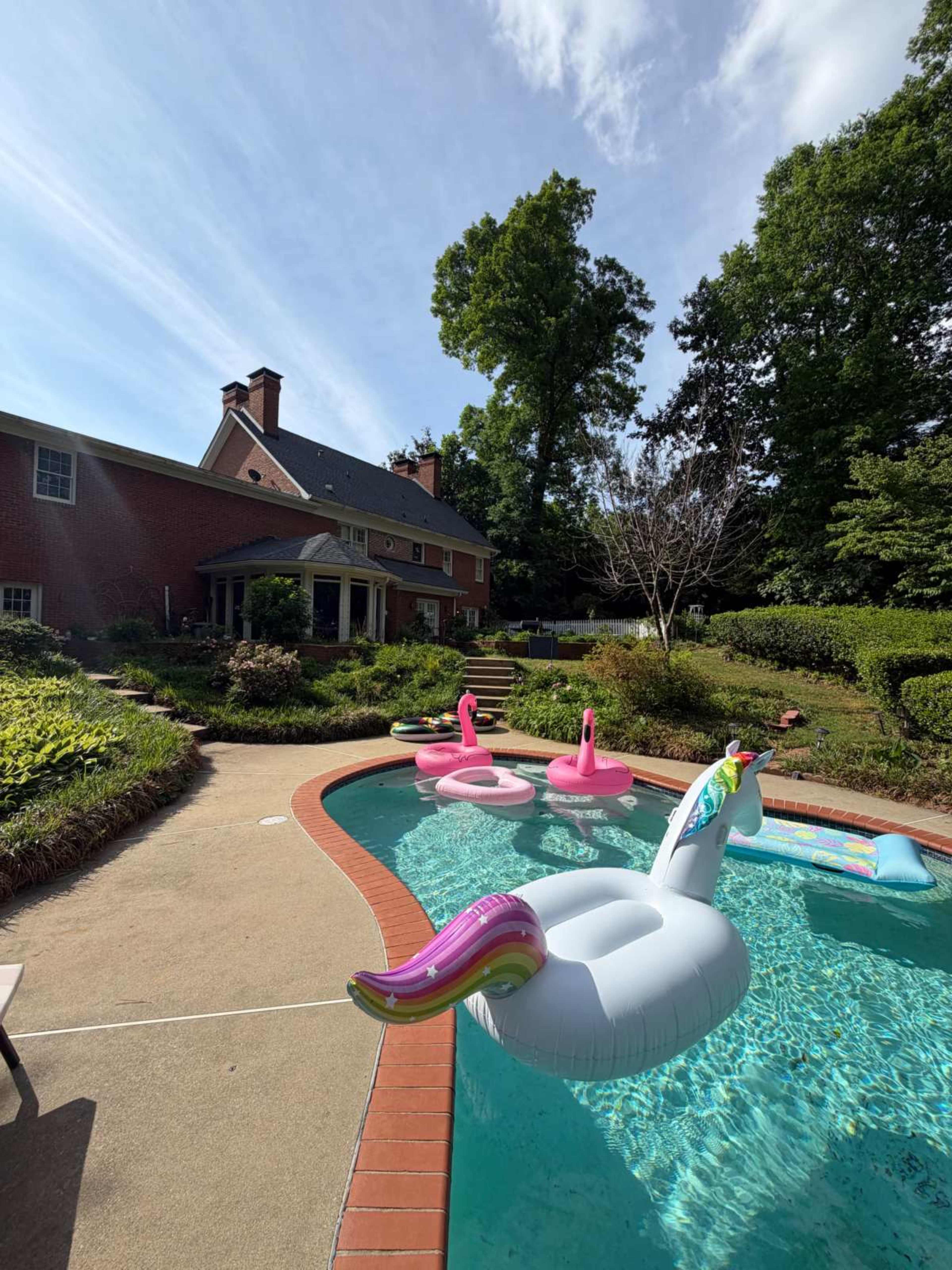 The image shows a backyard pool with colorful floaties, including a unicorn and two flamingos, surrounded by a landscaped yard and a brick house.