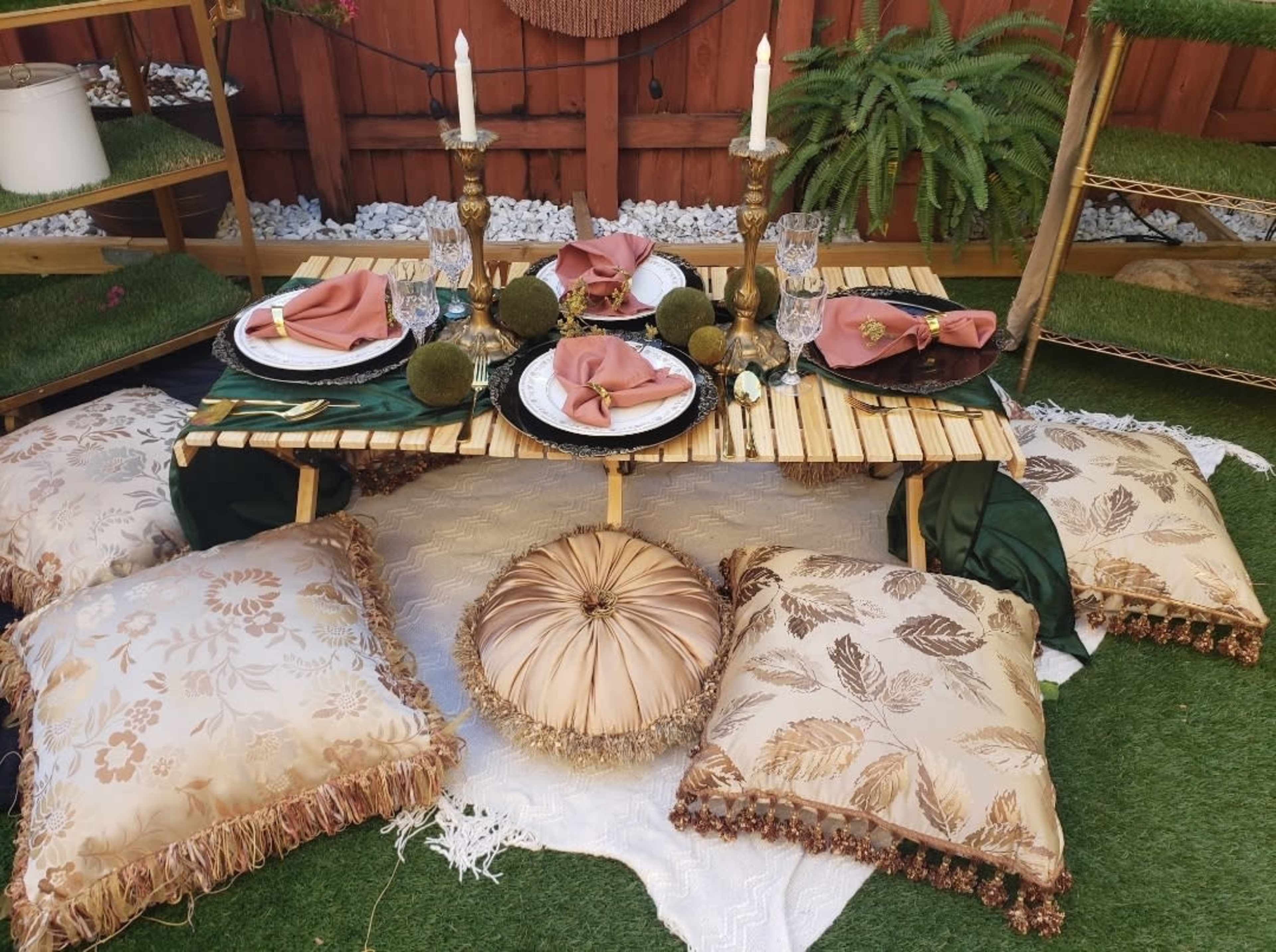 A low bamboo table set with dishes, napkins, and decorative items, surrounded by plush pillows on a green lawn.