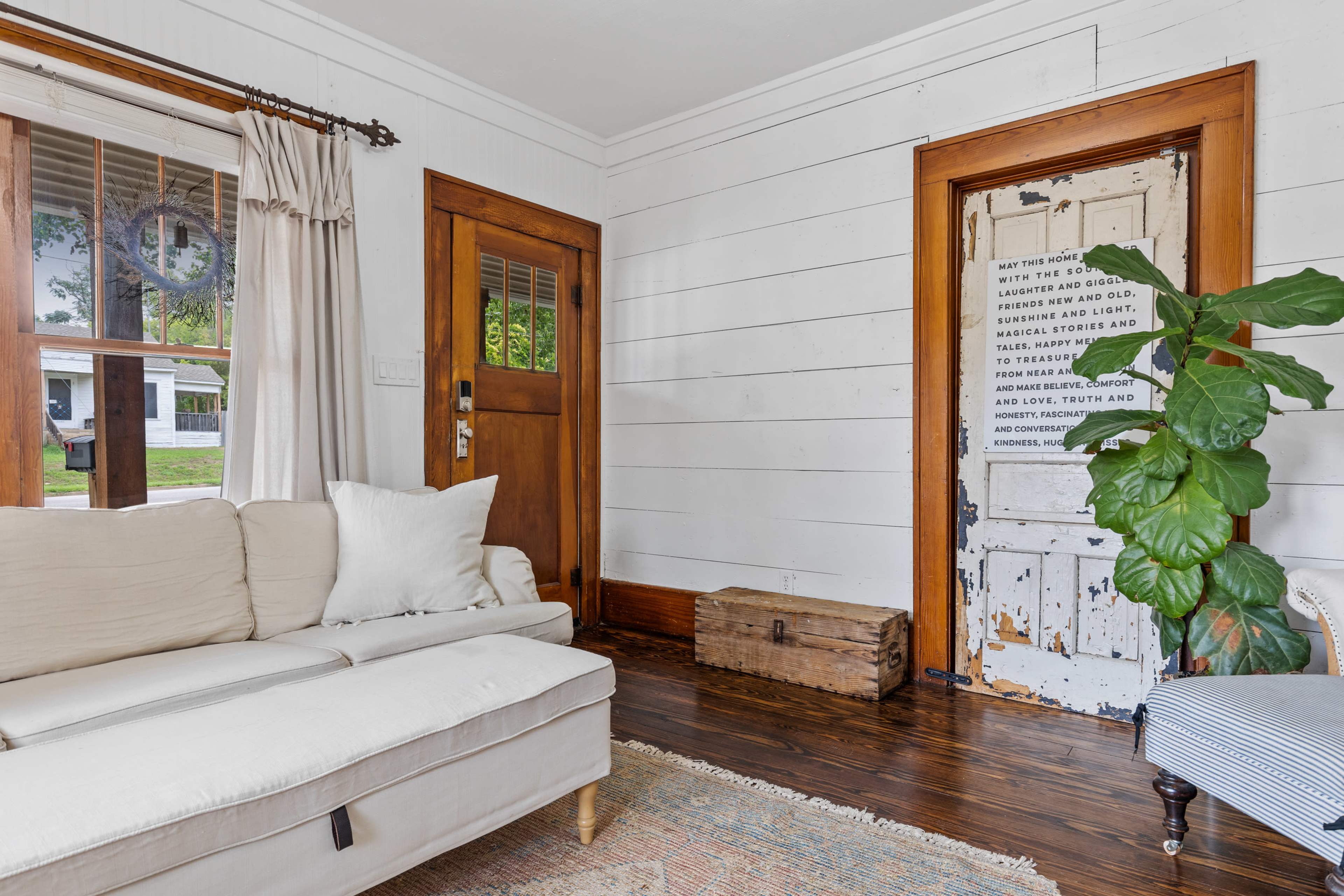 A living room with a light-colored sofa, a wooden door with text, and a potted plant beside a rustic chest on the floor.