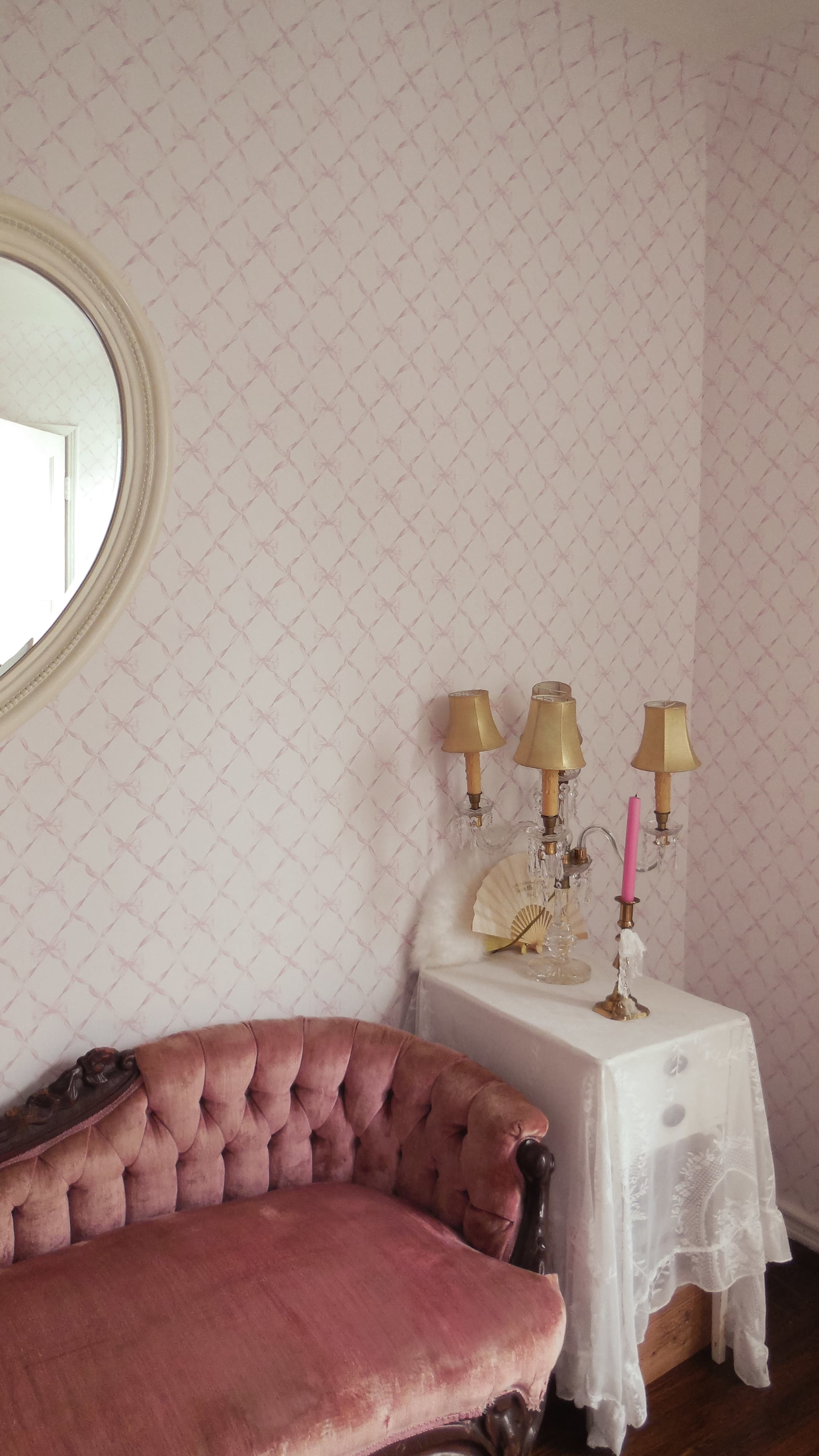 A pink sofa rests beside a table with two lamps and a decorative fan in a room featuring patterned wallpaper and a round mirror.