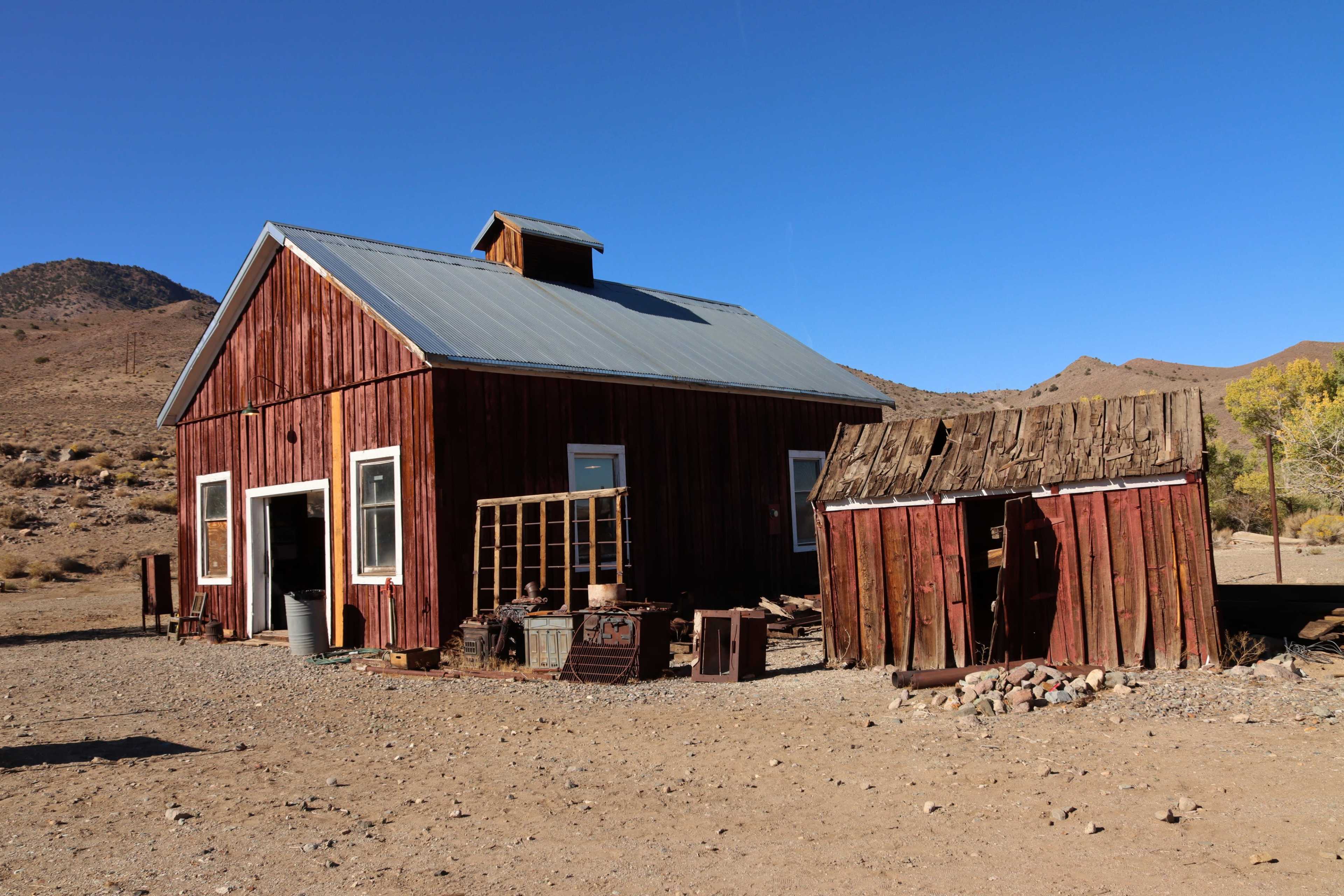 A red wooden house with a metal roof stands next to a smaller, weathered shed in a dry, desert landscape.