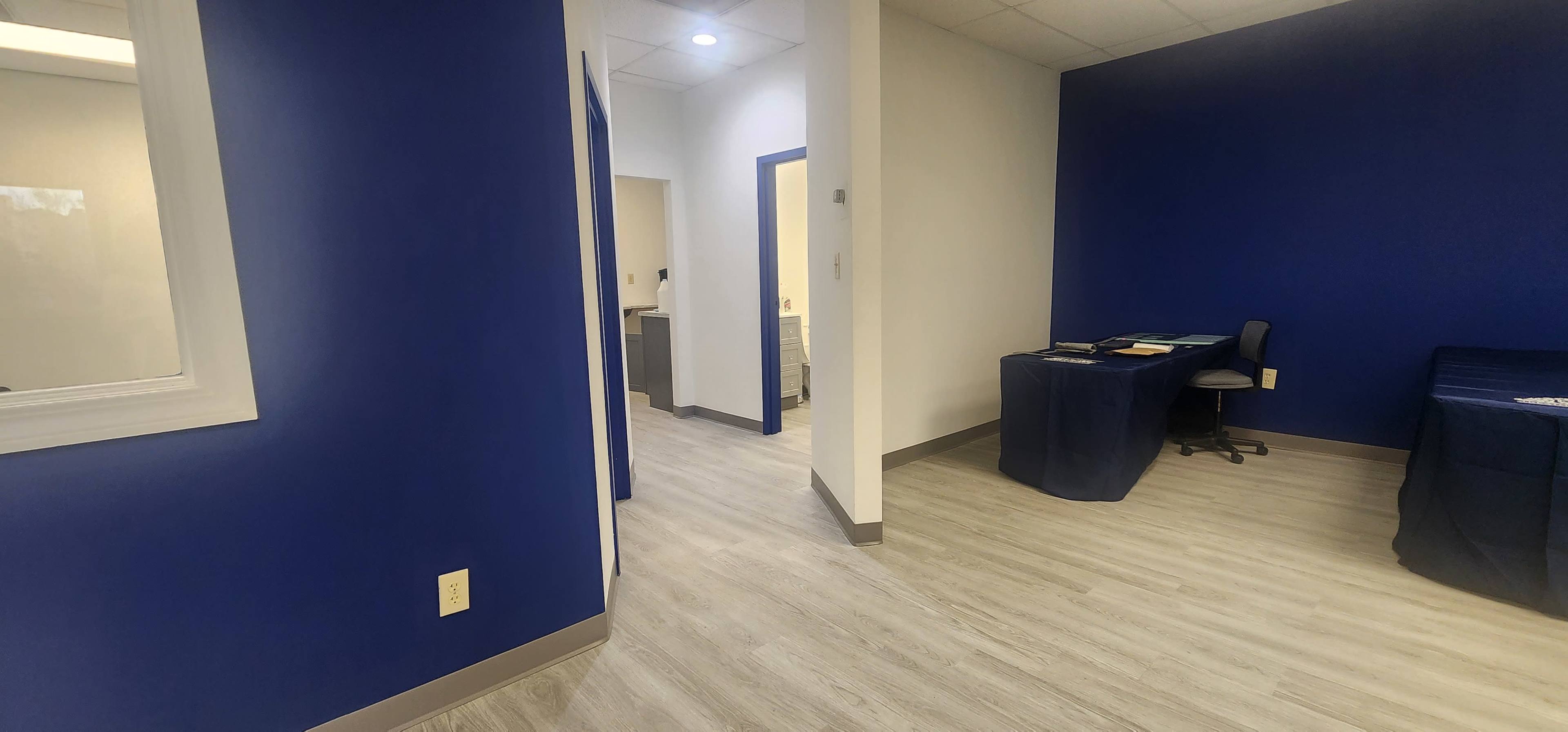 The image shows a brightly lit room with blue walls, wooden flooring, a small desk, and an open doorway leading to another area.